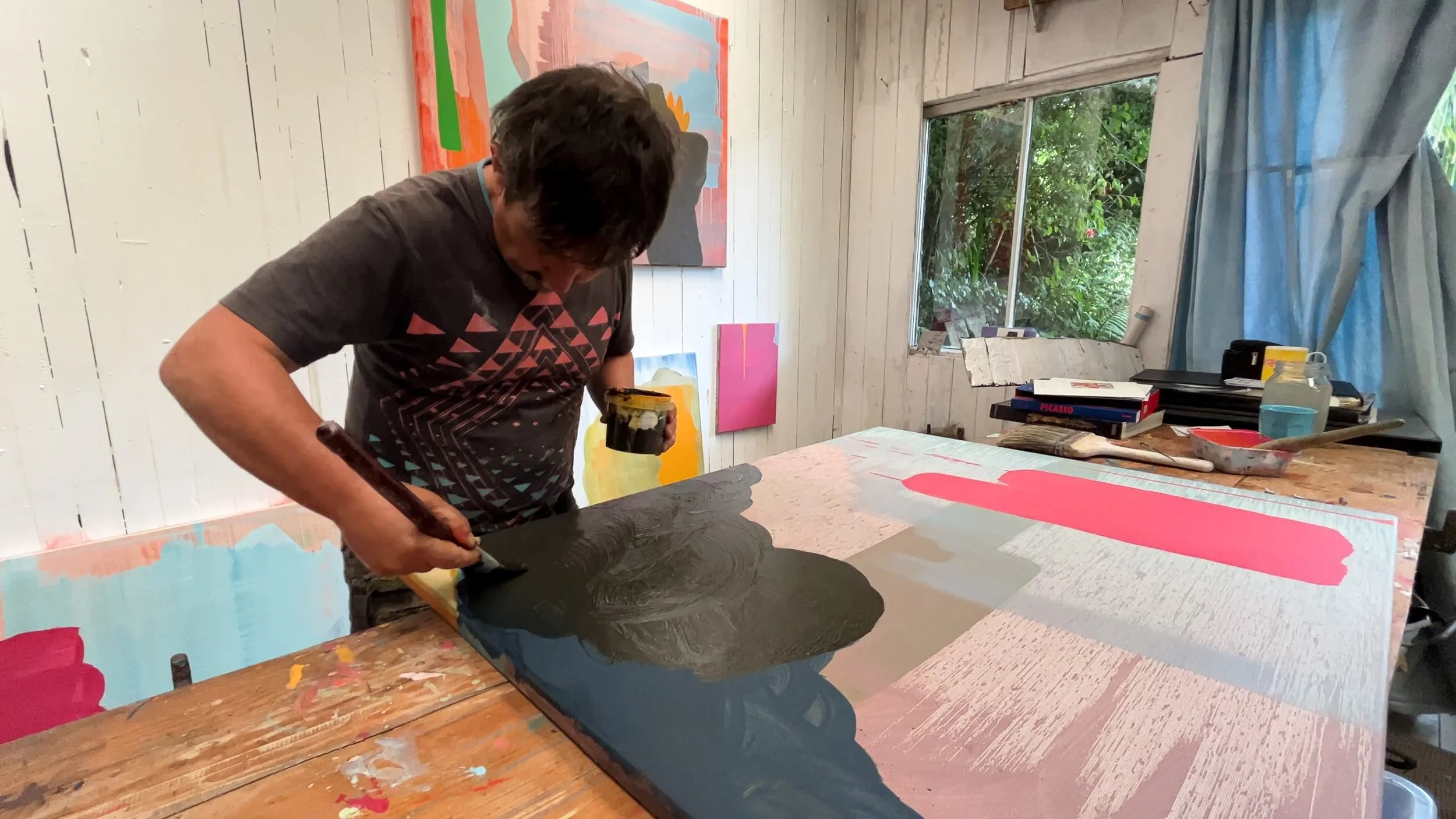 Irish artist Martin Finnin painting dark cloud form onto large abstract canvas on studio floor, pink and grey forms visible, multiple paintings in progress on white panelled studio wall behind, West Cork