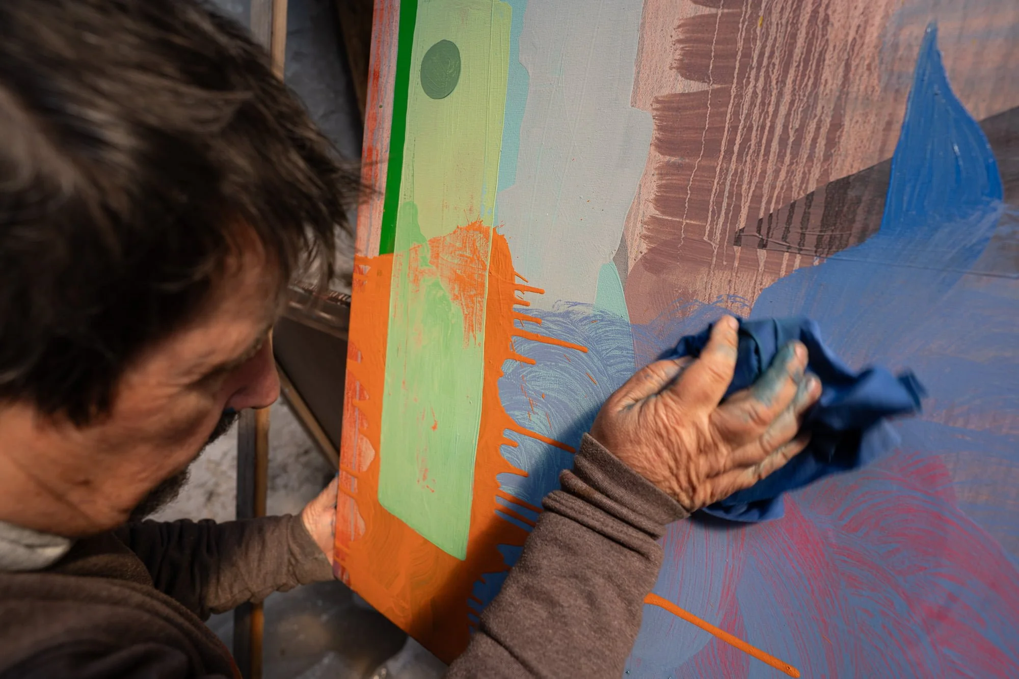 Close up of Irish artist Martin Finnin wiping blue paint with rag onto abstract canvas, vivid orange and green forms with drips, hands-on painting process in West Cork studio