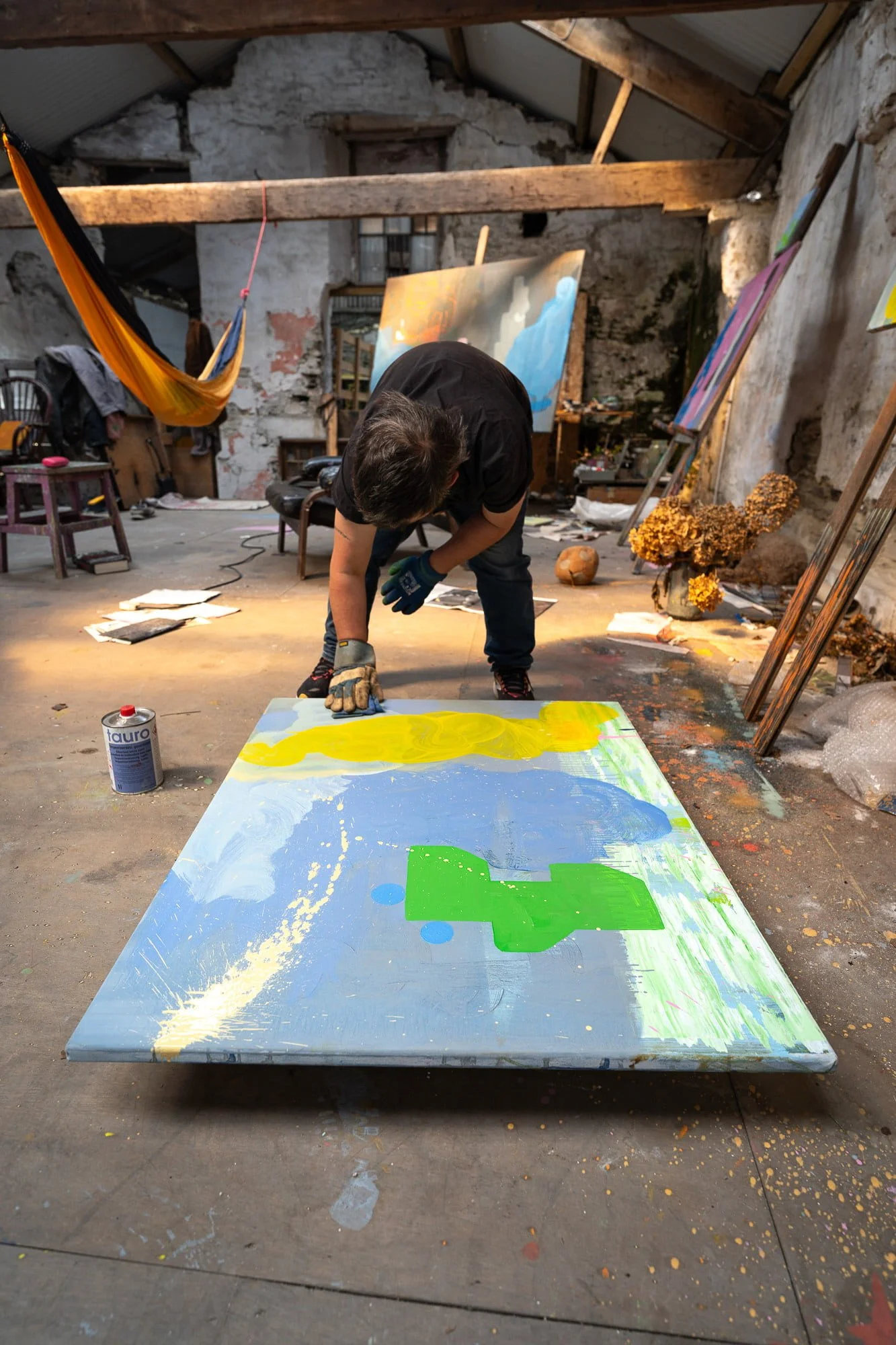 Irish artist Martin Finnin working on large abstract oil painting on studio floor, yellow and green forms emerging on canvas, artist studio with hammock and multiple canvases in background
