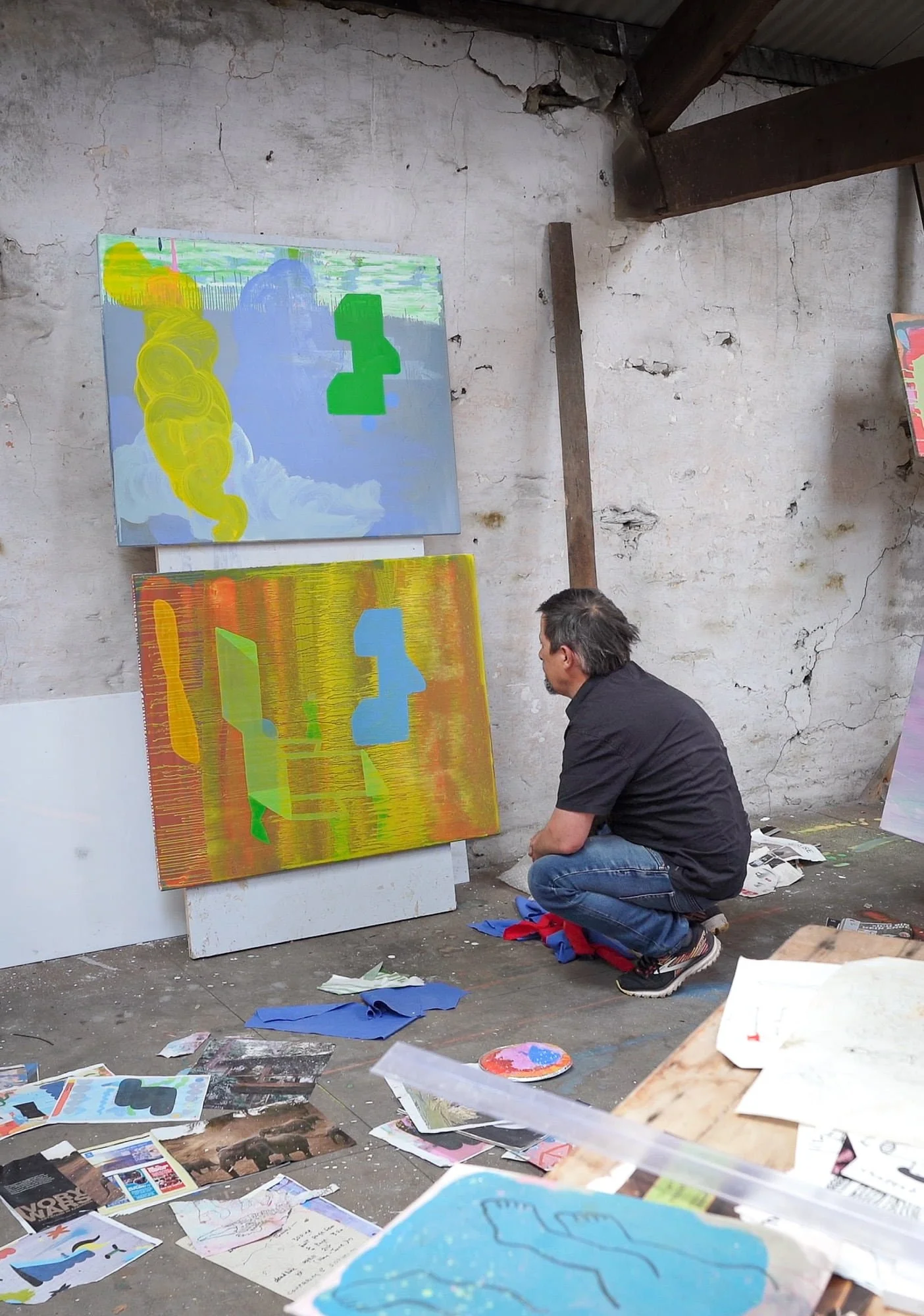 Irish artist crouching to study two abstract oil paintings in progress, yellow green blue and orange forms, West Cork studio with source material on floor, Martin Finnin
