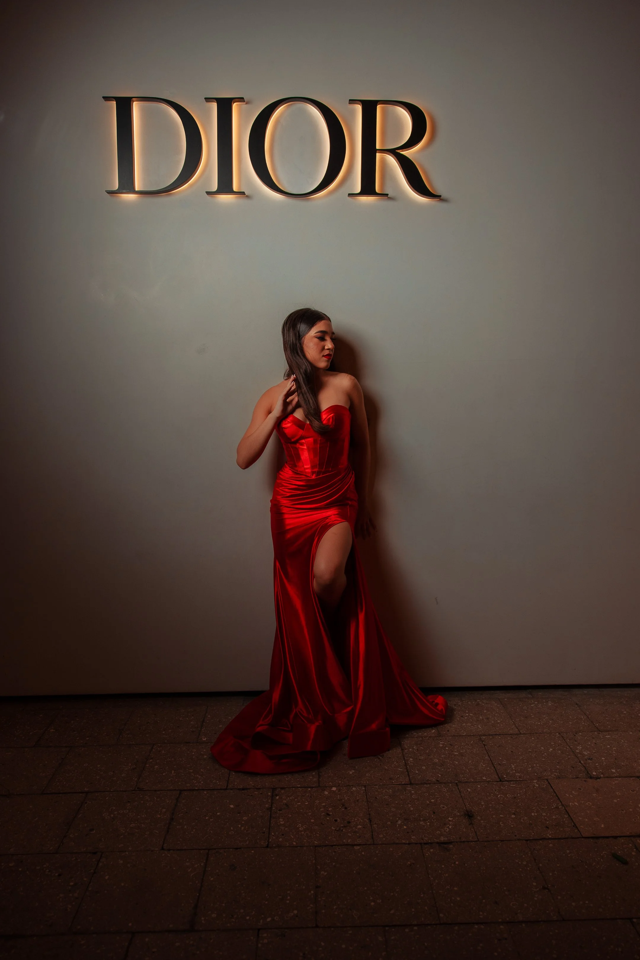 A woman in a red, satin, strapless gown with a high slit stands against a wall with the illuminated Dior logo above her.