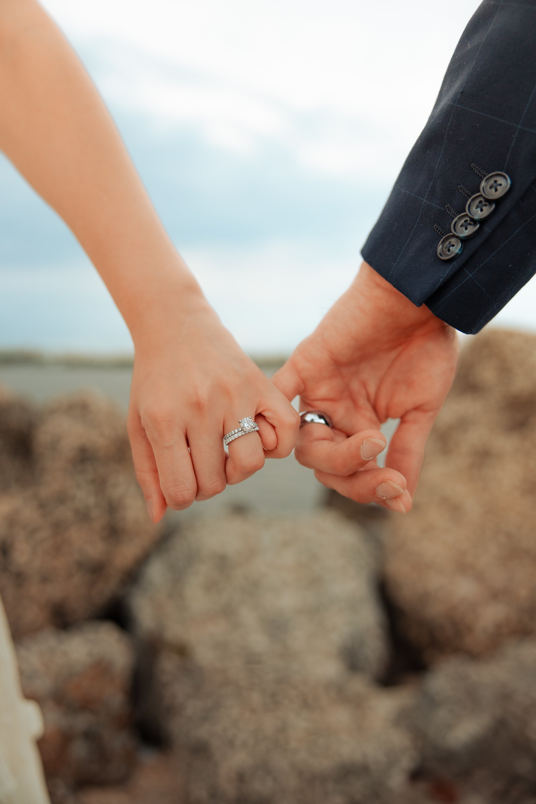 Close-up of a couple holding hands, showing wedding rings, with rocks and a cloudy sky in the background.