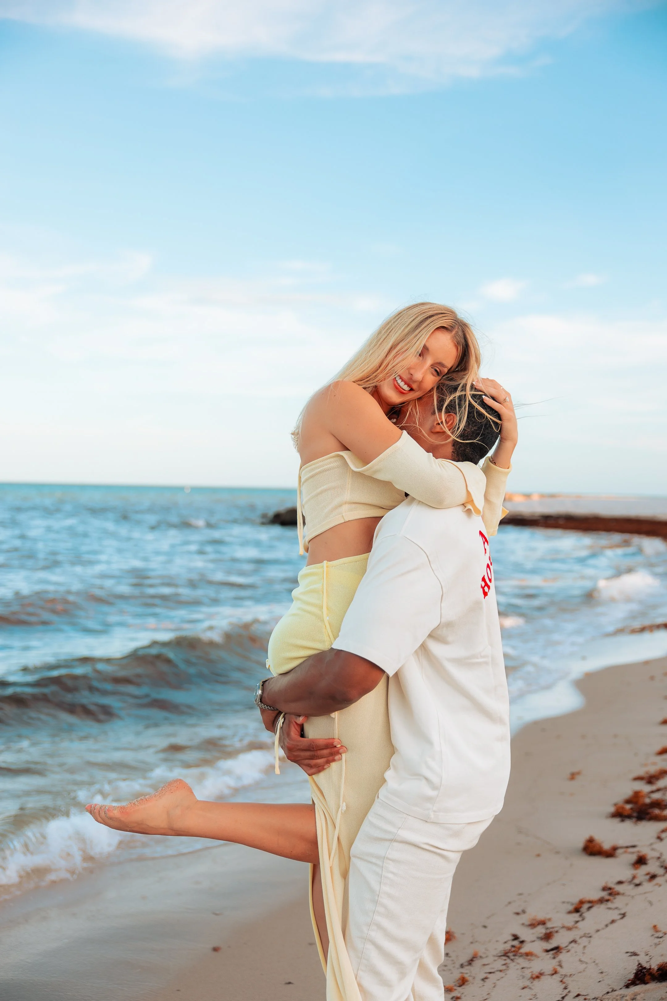 A couple embracing on the beach, with the man lifting the woman and both smiling, near the ocean with a blue sky.