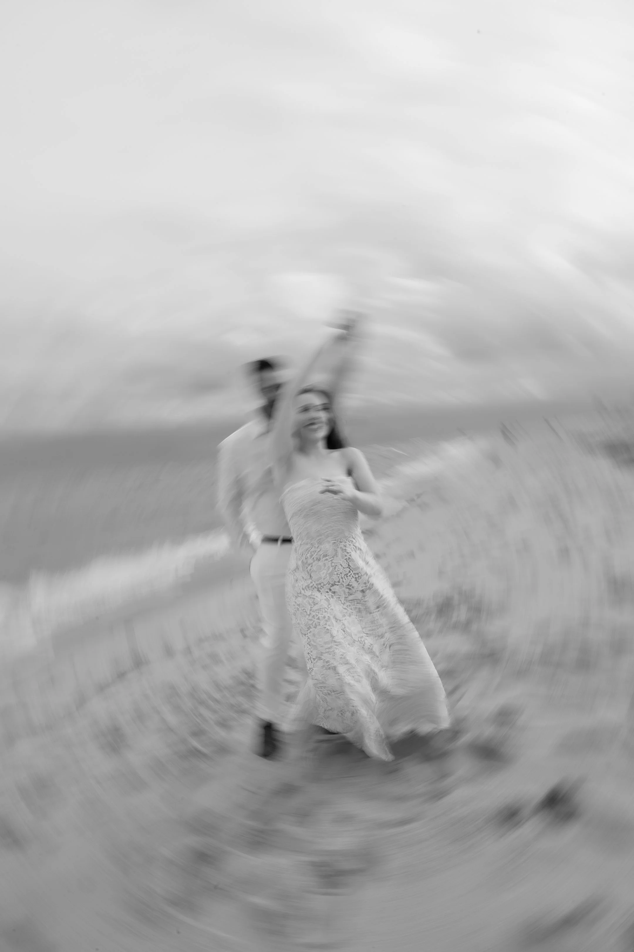 Black and white photo of a couple dancing on the beach, with a dreamy, blurred effect, and cloudy sky in the background.