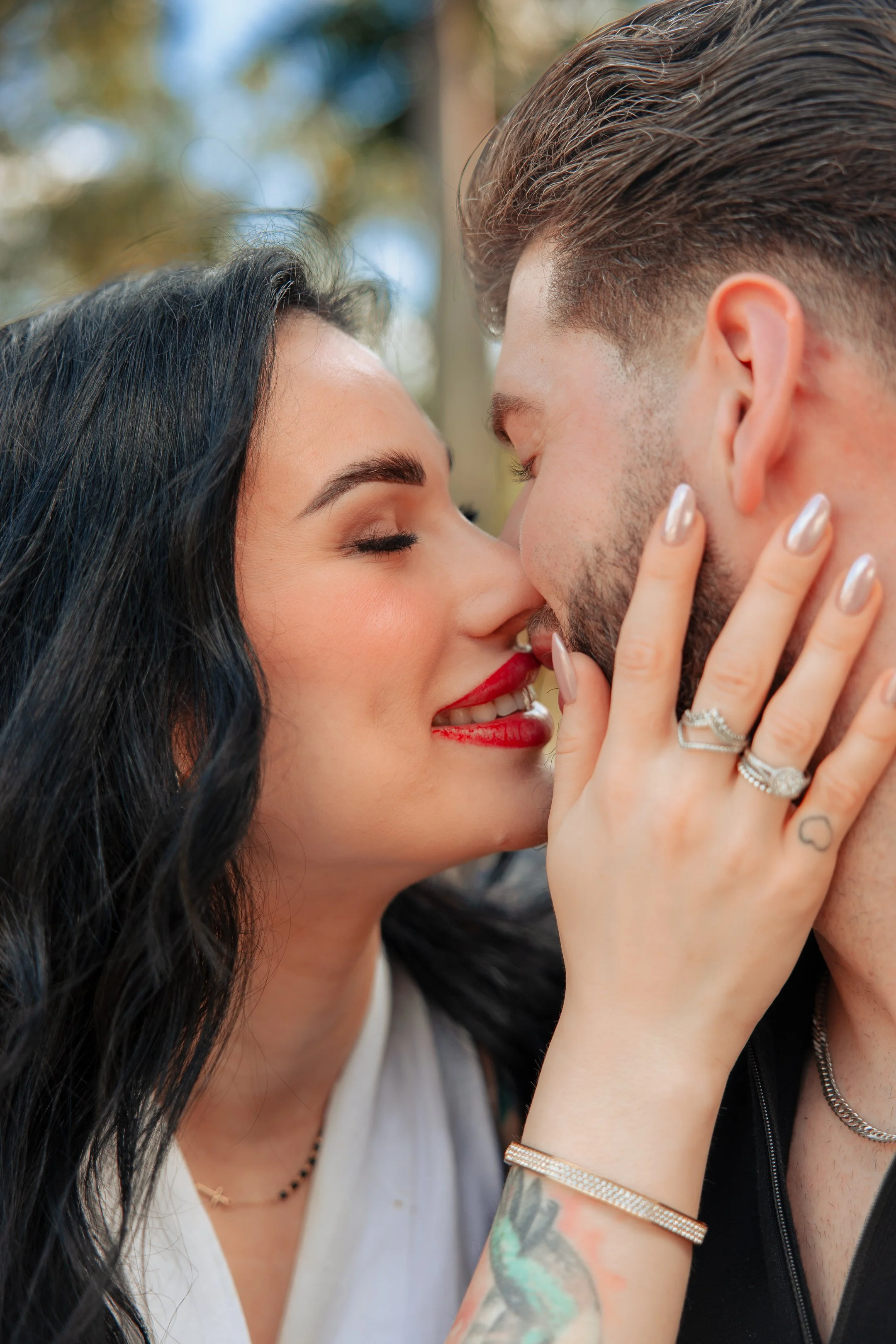 A woman and man kiss closely outdoors during daytime, with the woman's hand on the man's face, showing her rings and a tattoo on her arm.