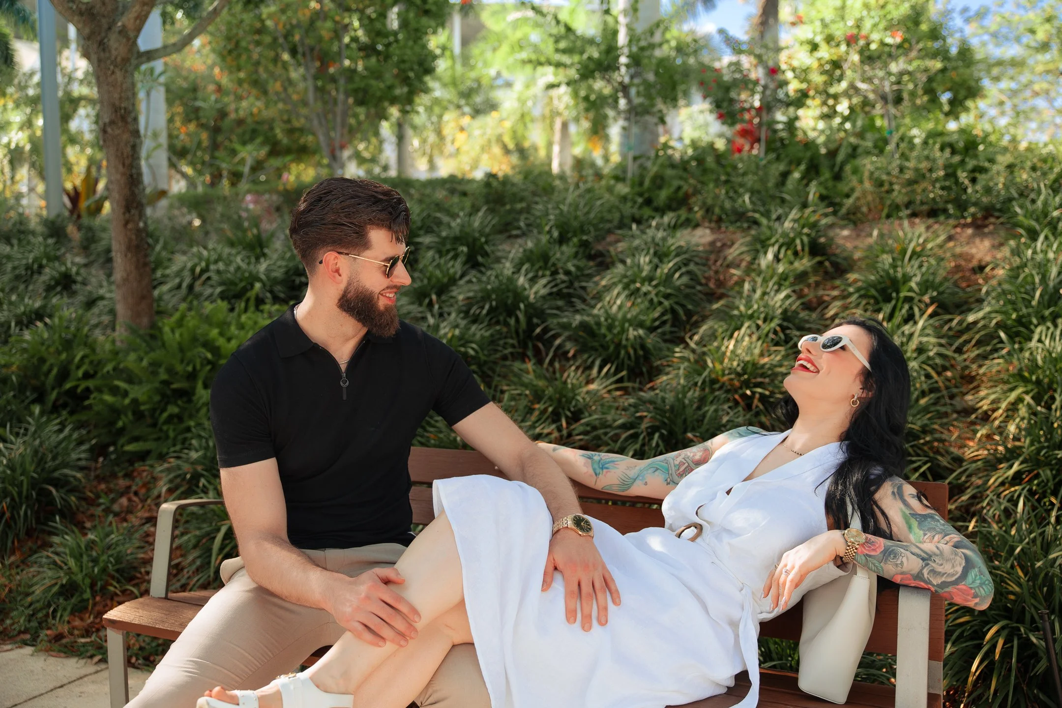 A man and a woman with tattoos and sunglasses are sitting on a park bench together outdoors. The woman is lying back, smiling, with her arm around the man, who is touching her knee. They are surrounded by green foliage and trees.
