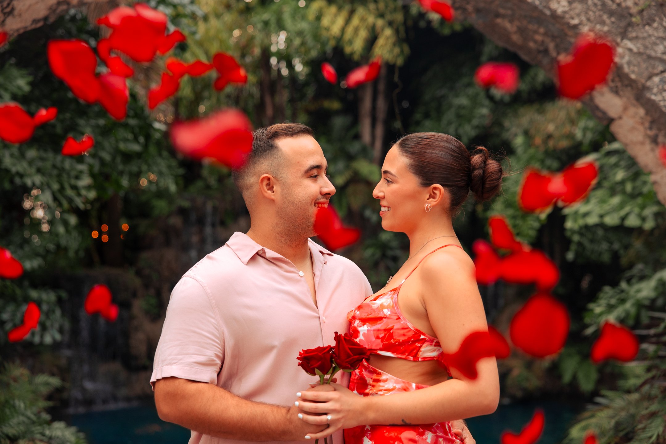 A couple smiling and looking at each other, with roses and petals falling around them outdoors in a lush green setting.