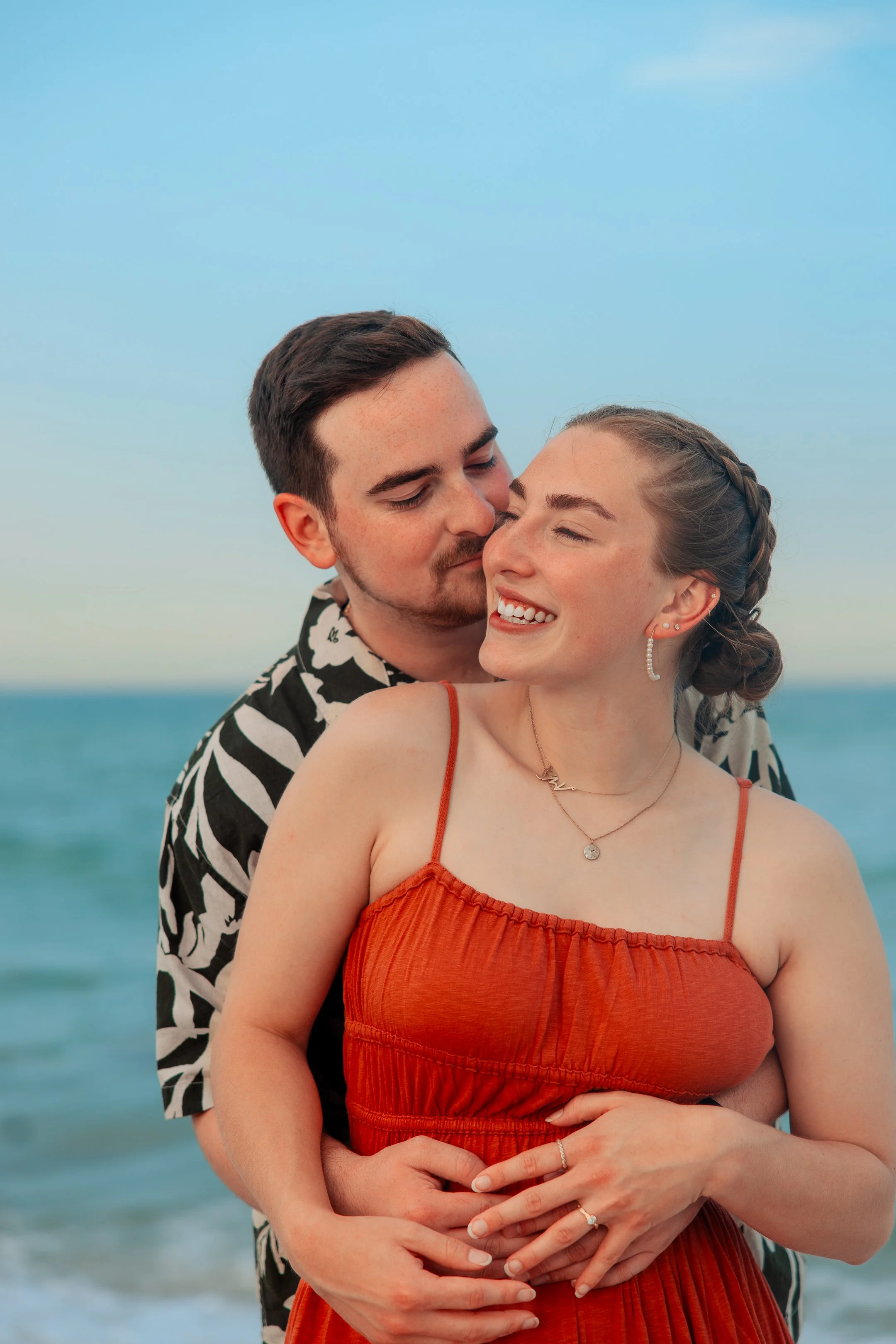 A couple happily embraces on a beach, with the man in a patterned shirt and the woman in an orange dress, both smiling with their eyes closed.