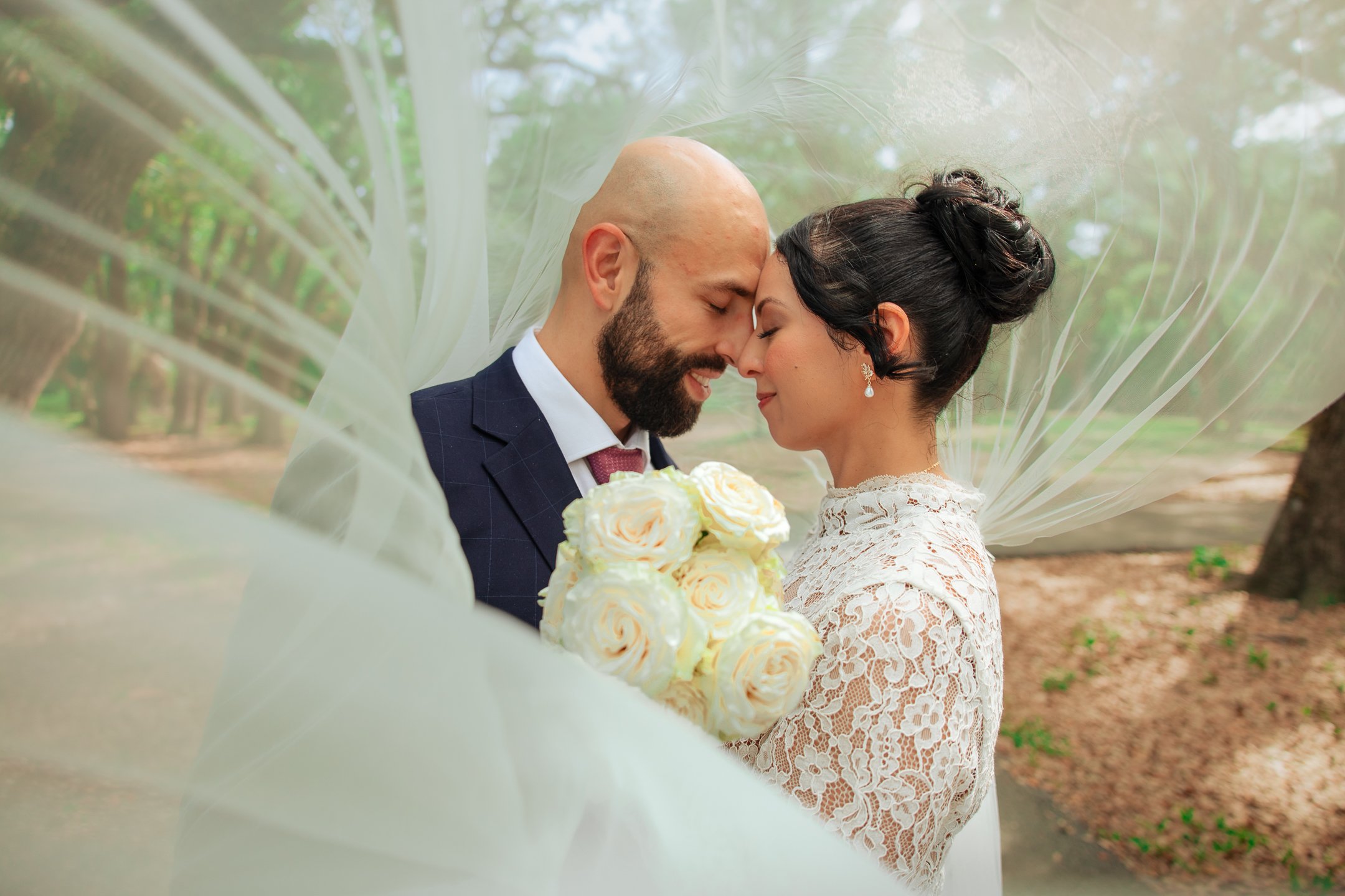 A bride and groom standing close together, with foreheads touching and eyes closed, in an outdoor setting. The bride wears a lace dress and earrings, holding a bouquet of white roses. The groom is dressed in a suit and tie, with a bald head and beard