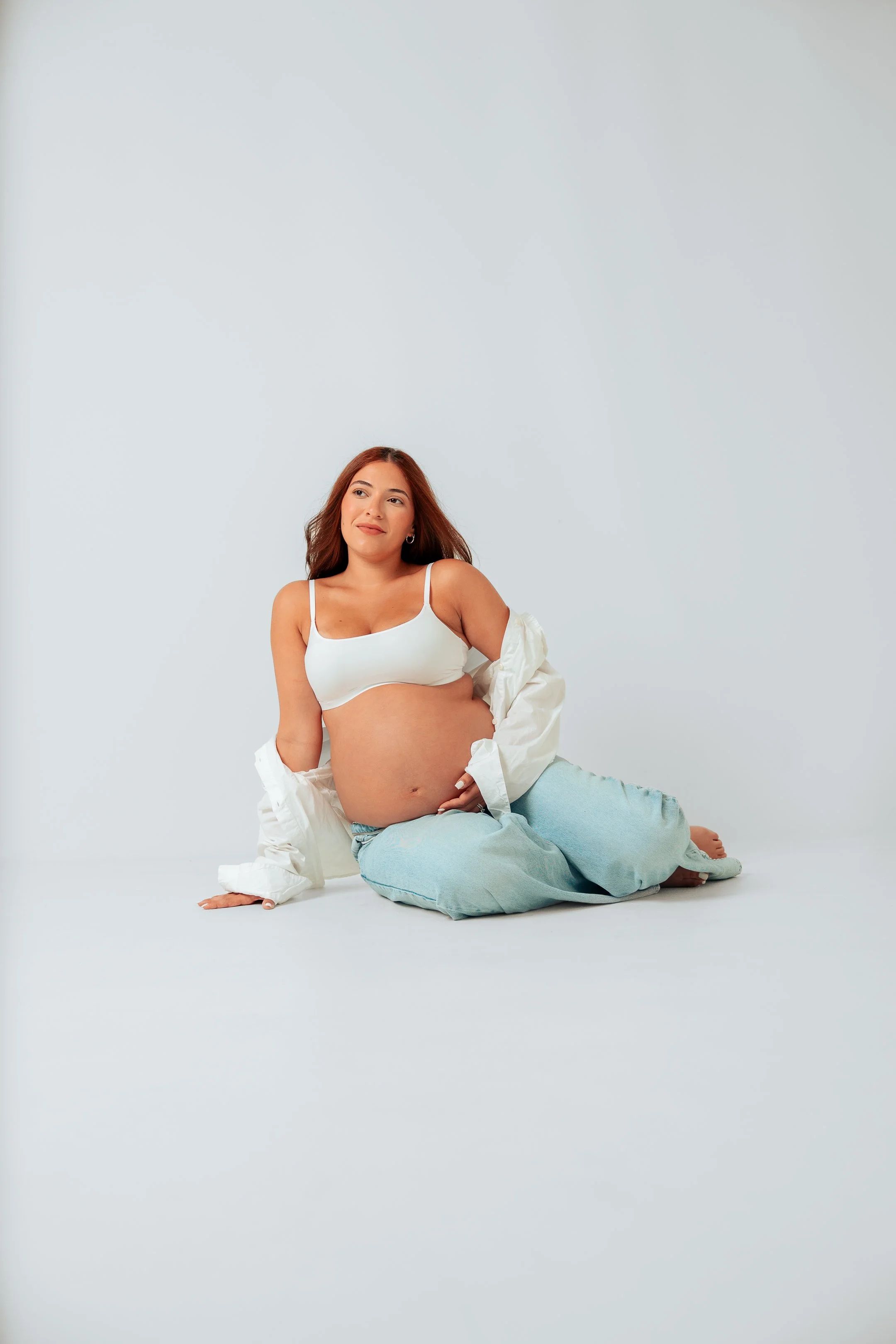 Pregnant woman with long brown hair sitting on the floor in a white tank top, light blue pants, and an open white shirt against a plain light background.