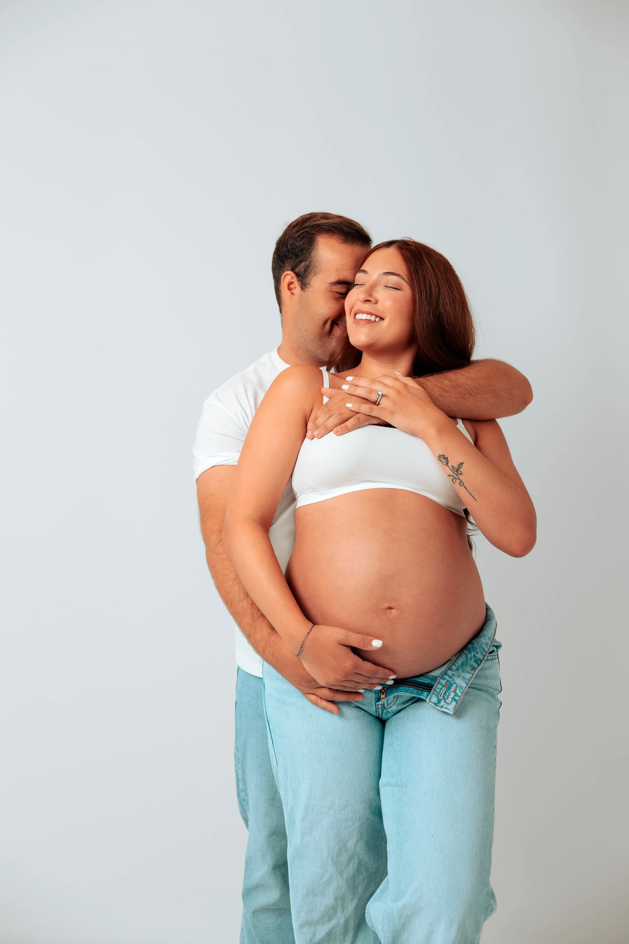 A pregnant woman with red hair in a white crop top and light blue jeans, hugging a man from behind, both smiling with eyes closed, against a plain gray background.