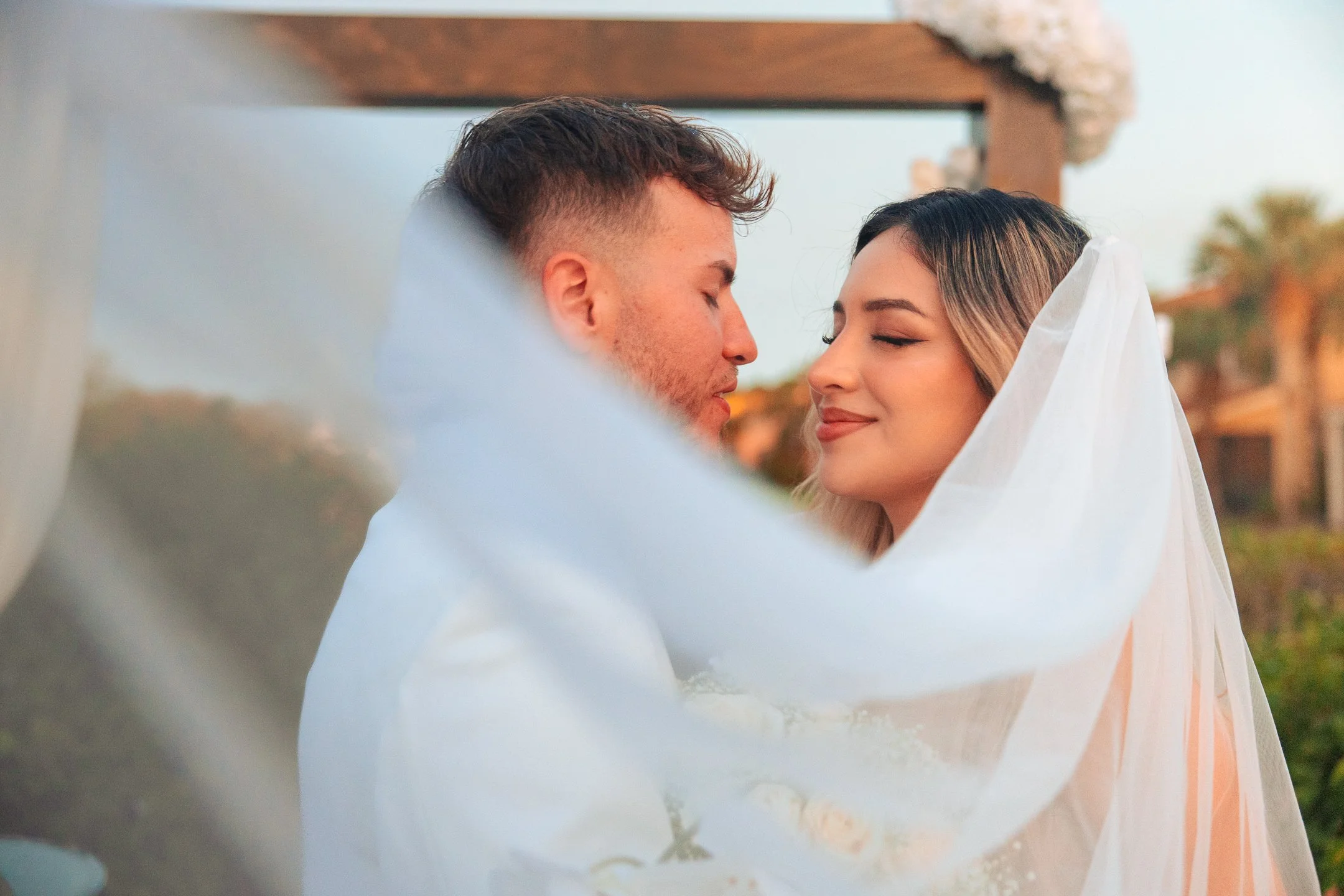 A bride and groom sharing a kiss, with the bride wearing a wedding veil and the couple outdoors at sunset.