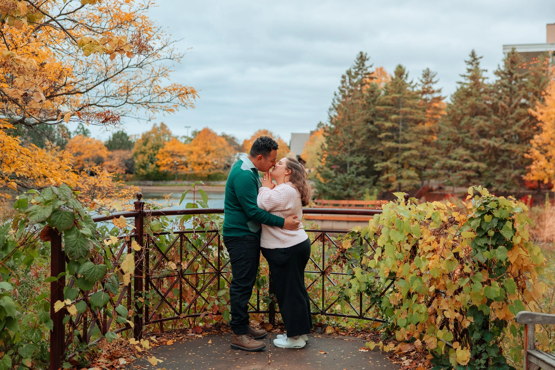 A couple sharing a kiss on a bridge surrounded by autumn foliage with orange and green leaves during daytime.
