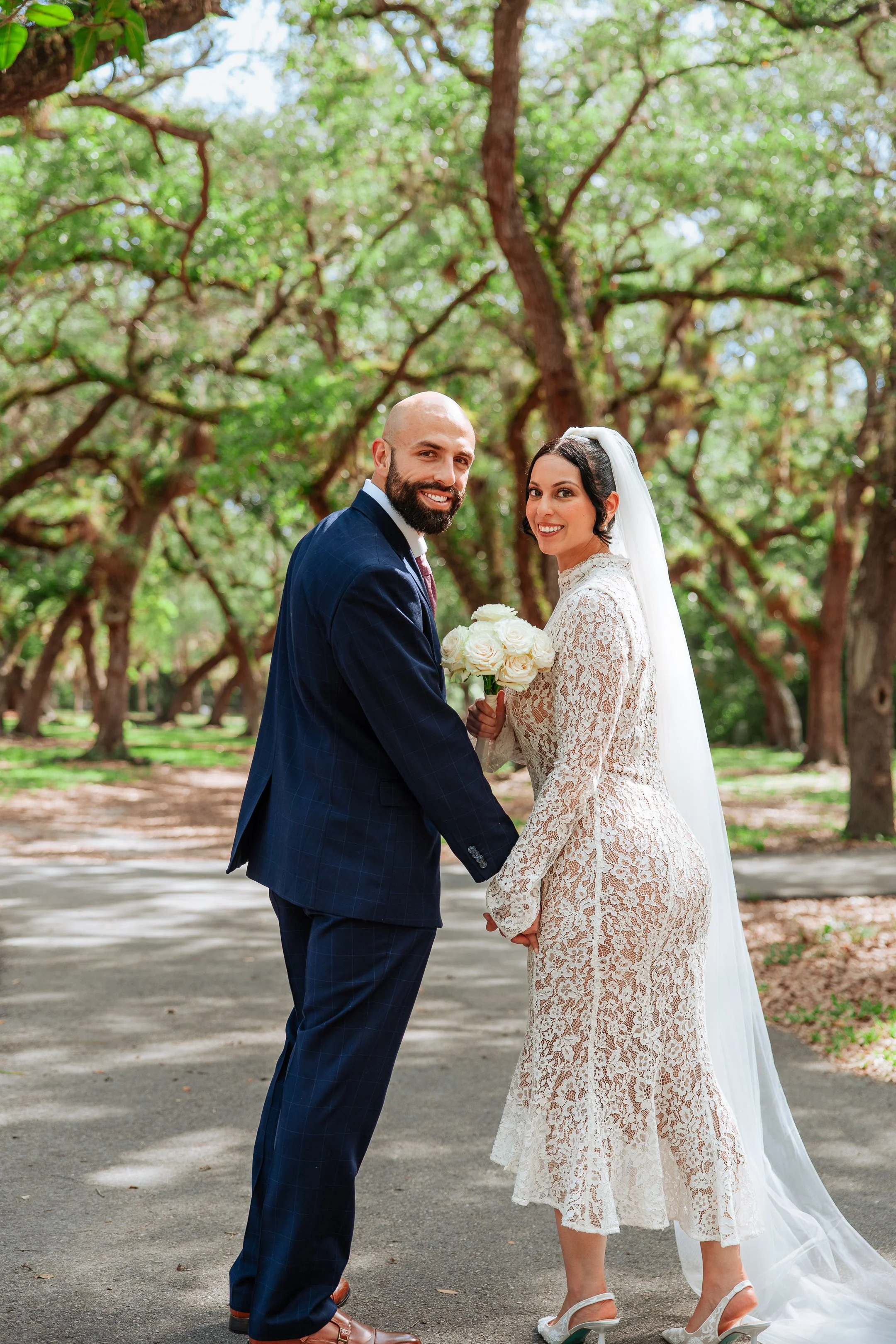 A newlywed couple holding hands and smiling in a park with green trees. The bride wears a lace wedding dress and veil, holding a bouquet of white roses. The groom wears a dark blue suit with a checkered pattern.