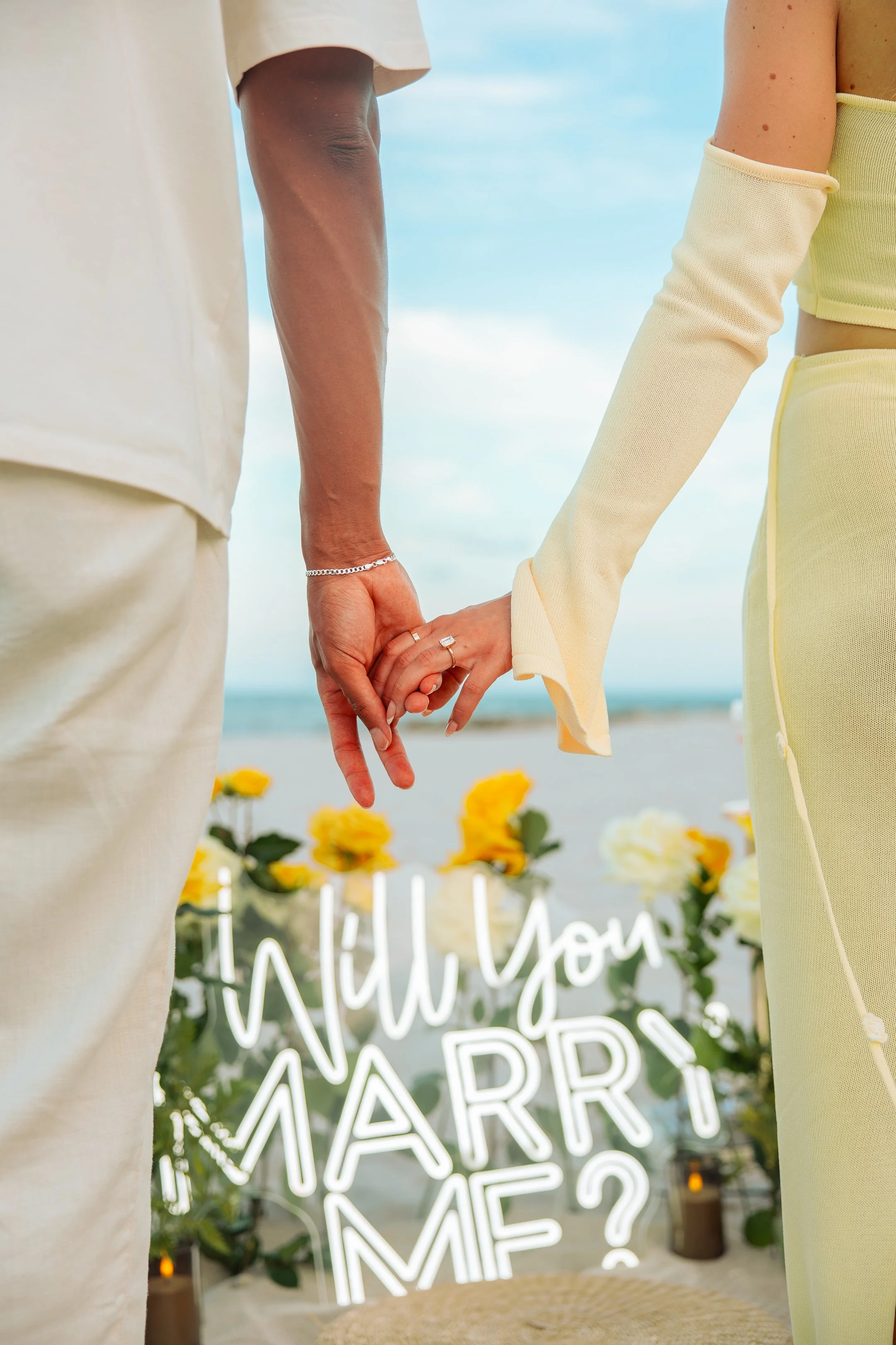 A couple holding hands titled 'Will You Marry Me?' on the beach with yellow flowers and candles in the background.