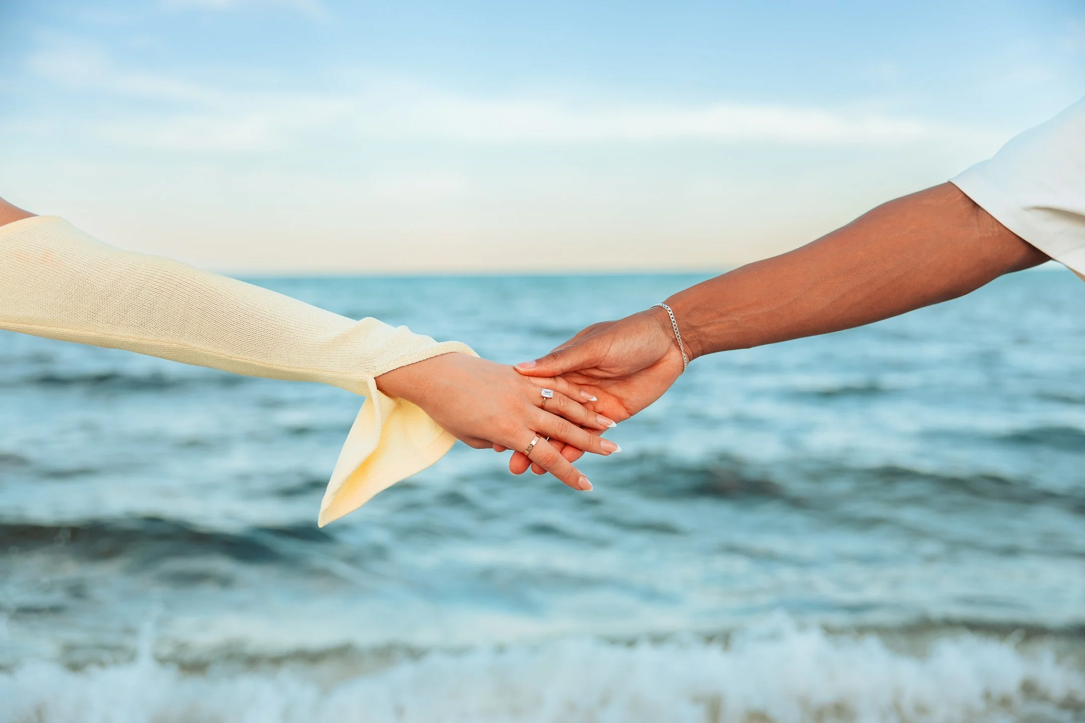 Two people holding hands near the beach with ocean waves and a clear sky in the background.