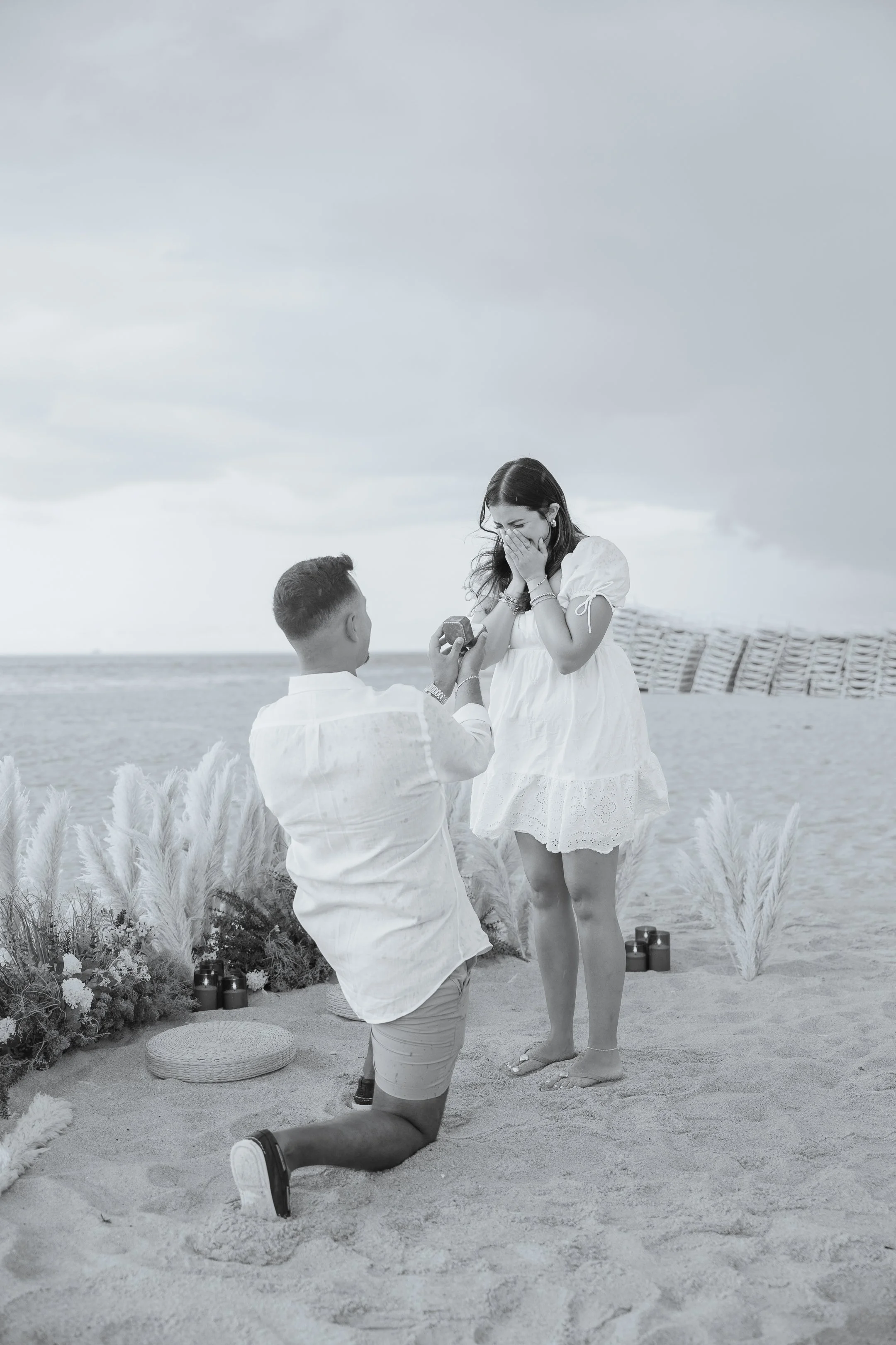 A man kneeling on the sand presenting a ring to a woman on one knee outdoors on a beach, with decorative plants and candles around them, during what appears to be a marriage proposal.