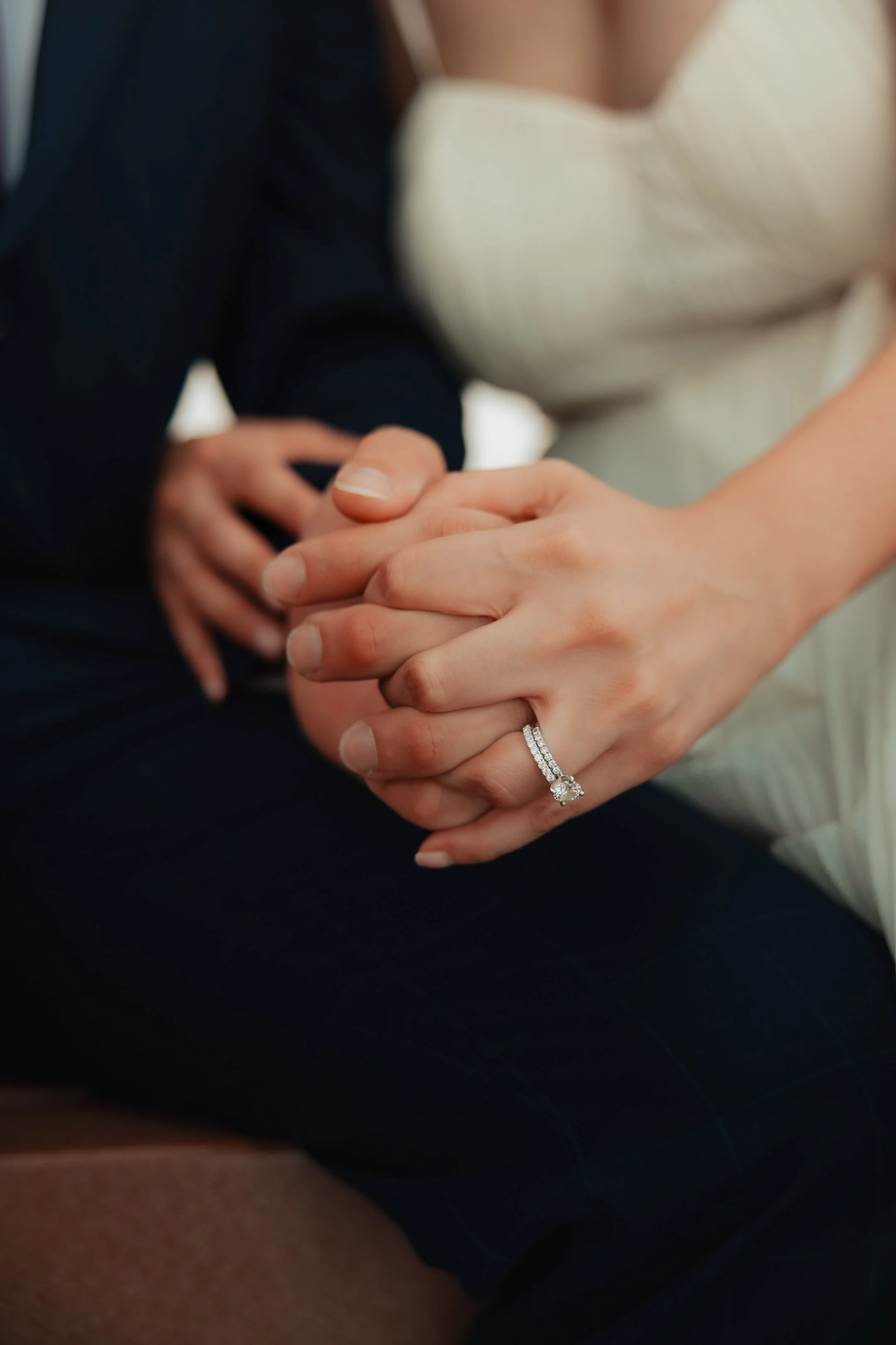 Close-up of a woman wearing a wedding ring, holding hands with another person, with both hands resting on her lap.