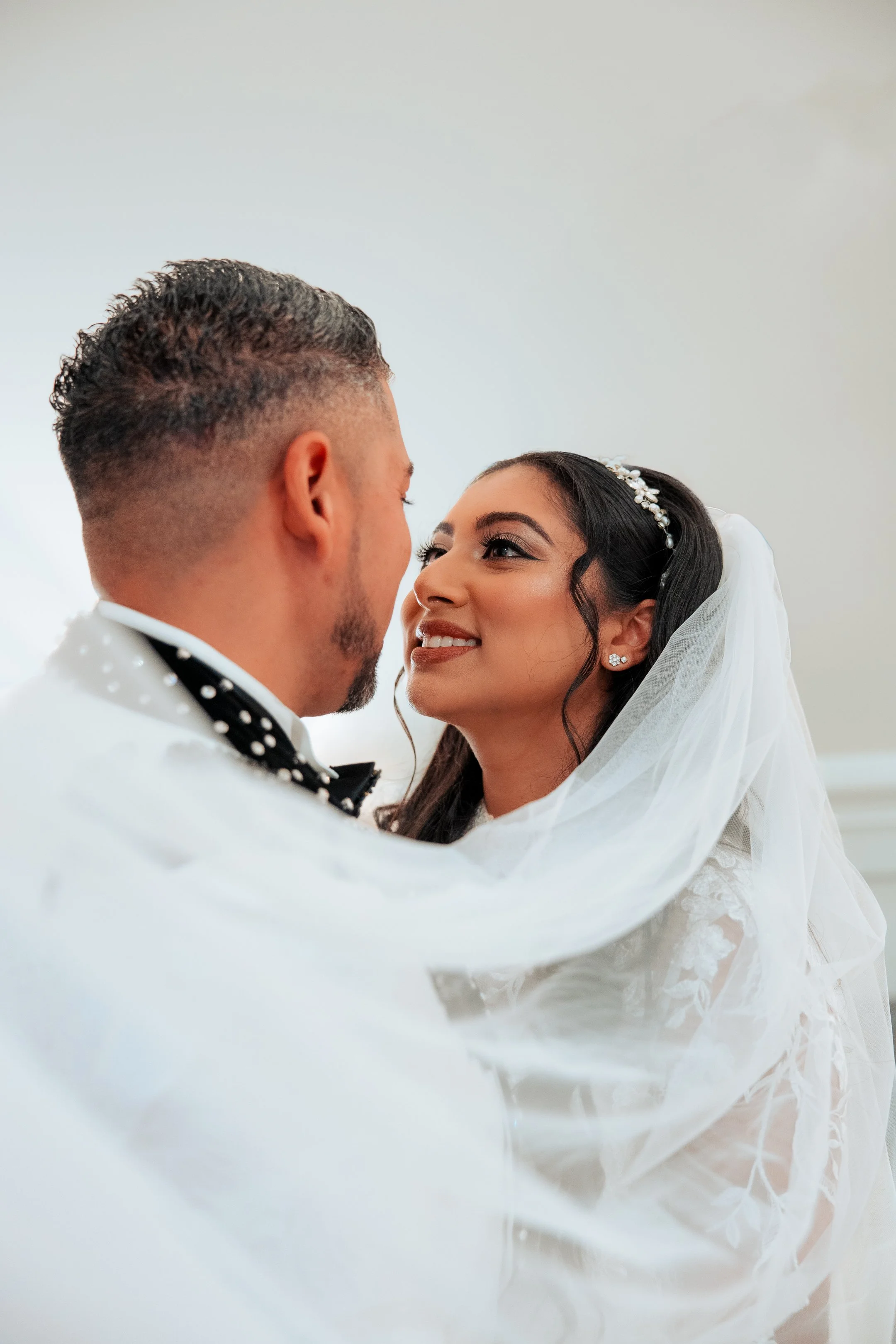 A bride and groom sharing an intimate moment during their wedding, looking into each other's eyes, with the bride smiling and wearing a white wedding gown and veil, and the groom in a tuxedo with a black bow tie.