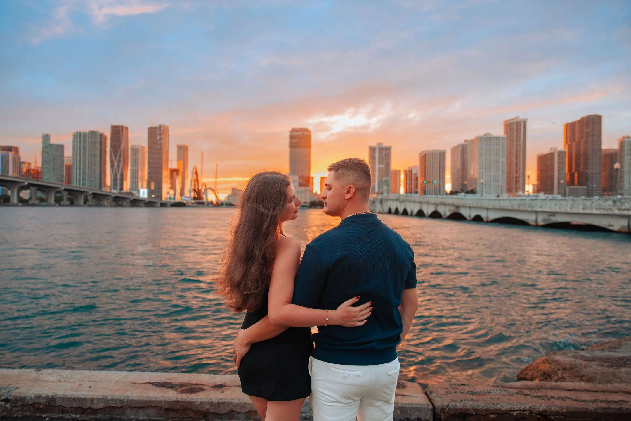 A couple standing by the water during sunset in a city with tall buildings.
