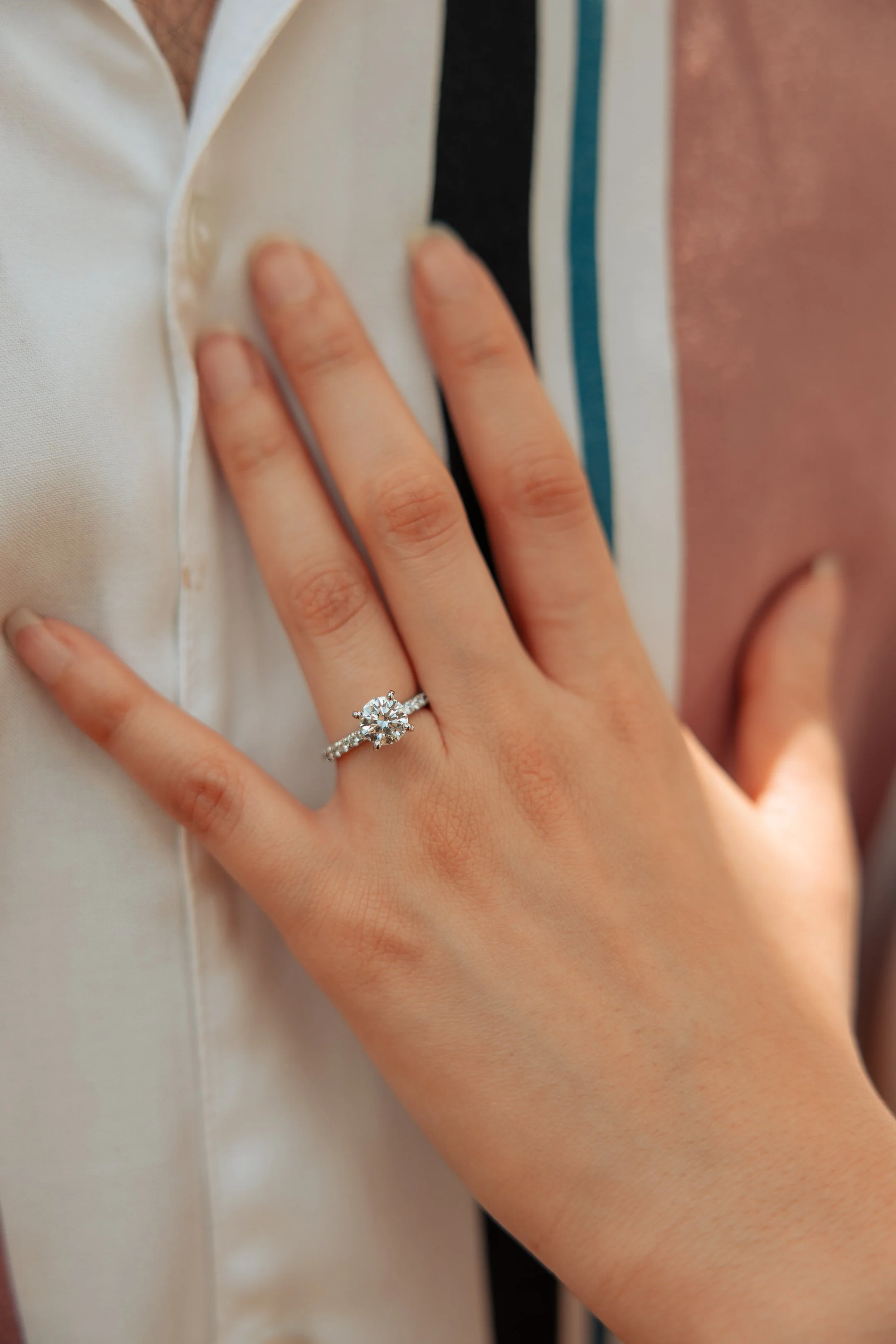 Close-up of a woman's hand wearing an engagement ring with a large central diamond surrounded by smaller diamonds, resting on a man's chest.