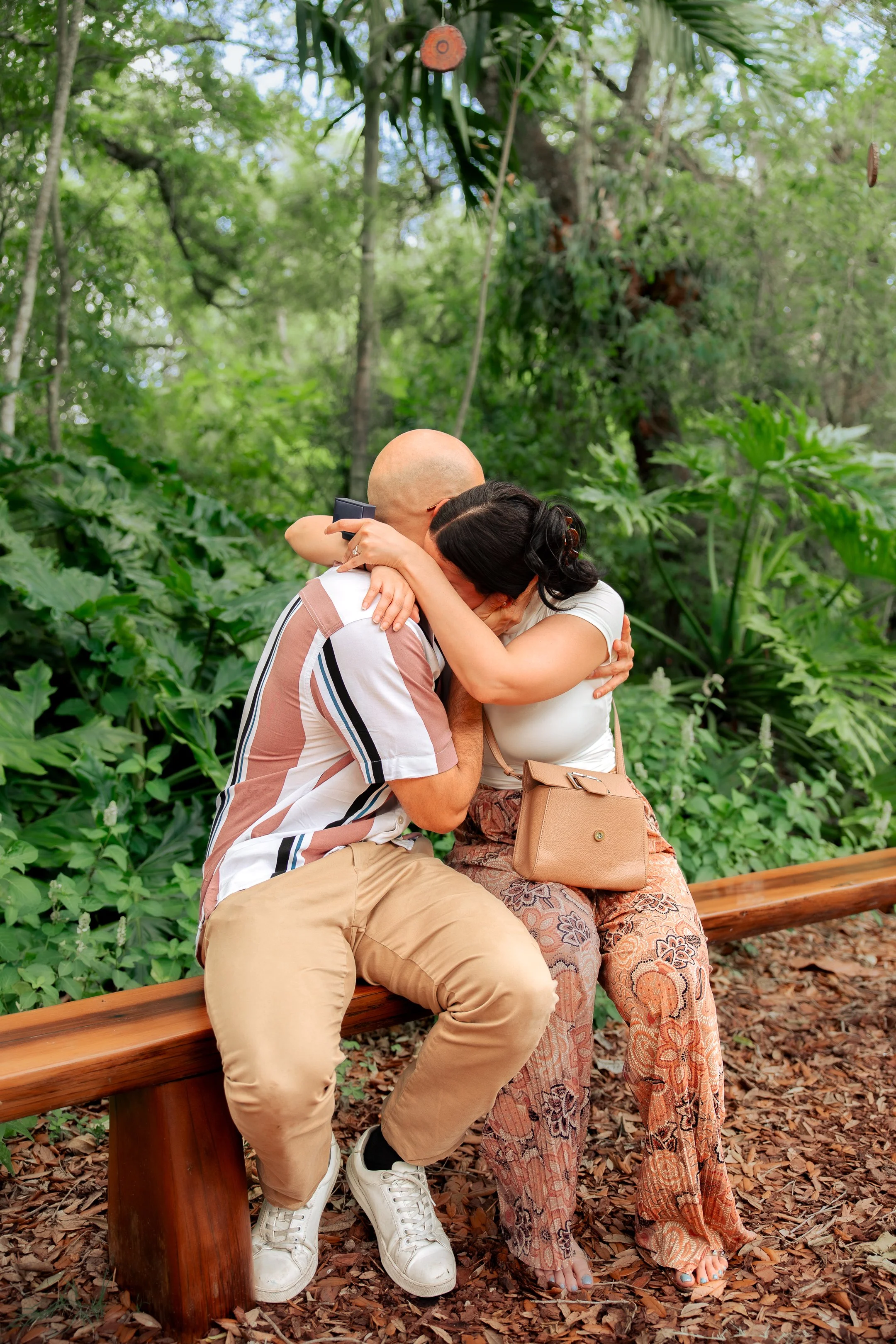 Two people sitting on a wooden park bench, embracing each other. One has a bald head and is wearing a short-sleeved shirt and beige pants. The other has black hair and is wearing patterned pants and a white top. They are surrounded by green foliage and tall trees.