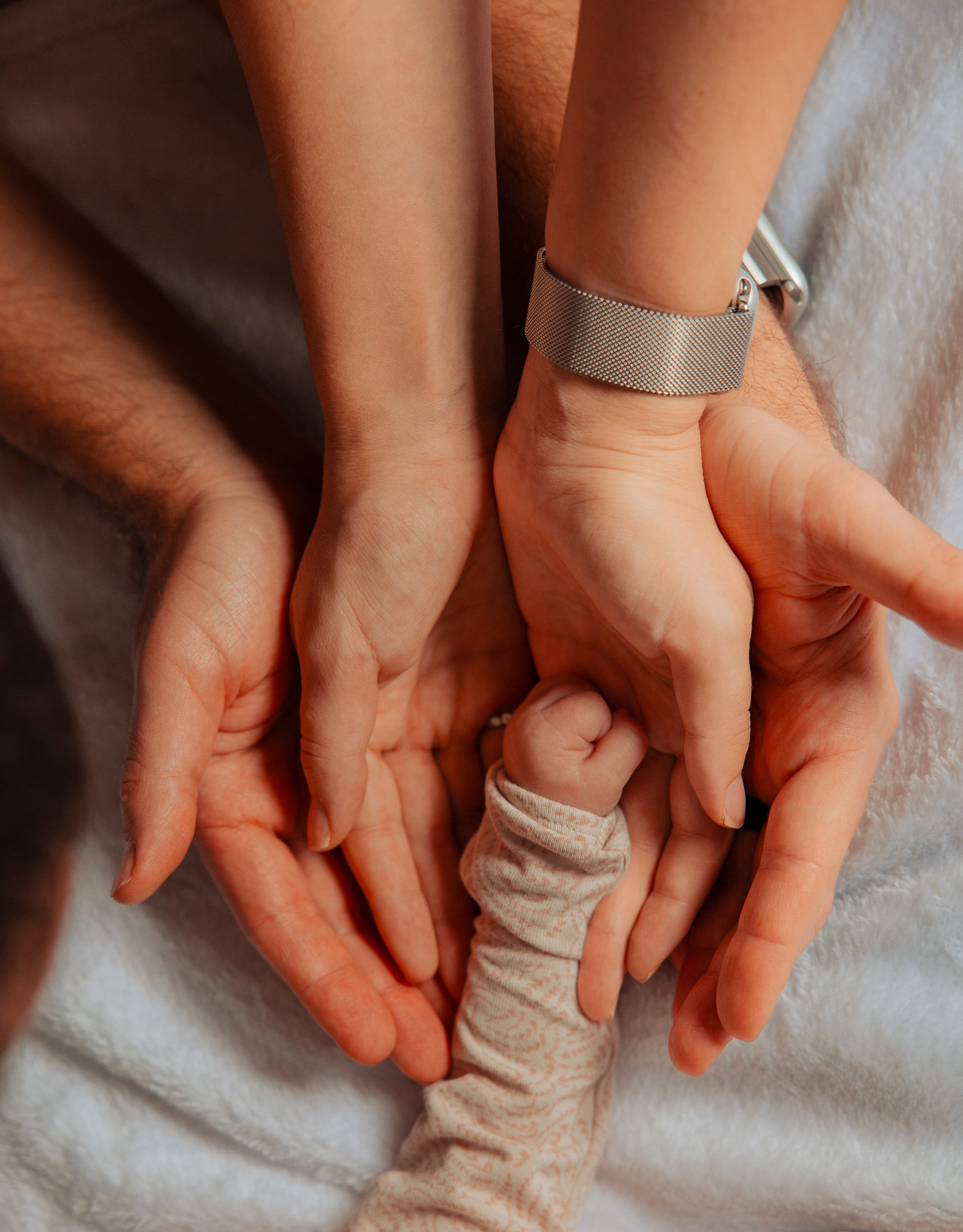 Close-up of an adult and child holding hands, showing the child's tiny hand clutching the adult's finger. The adult is wearing a silver watch.