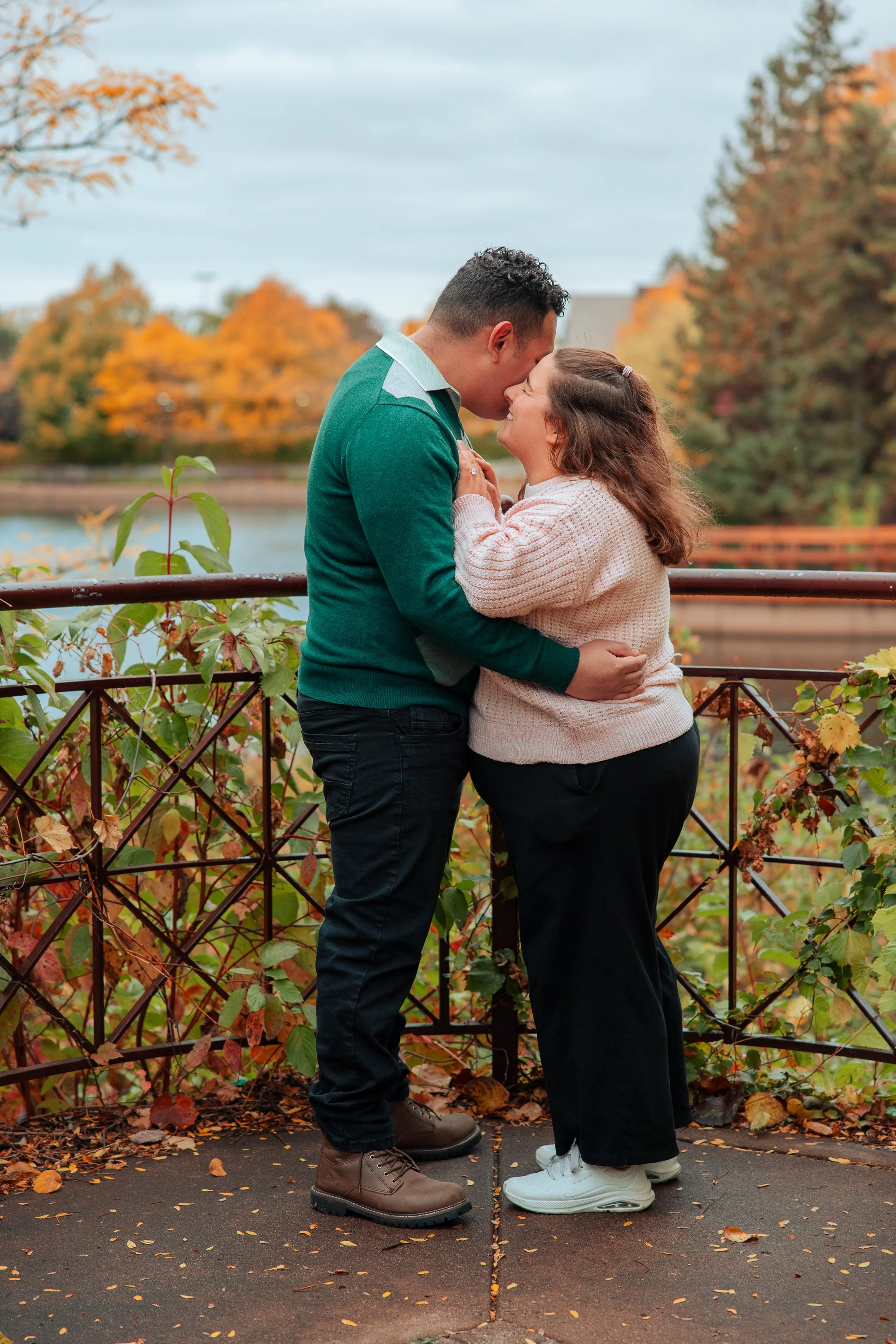 A couple in warm clothing shares a kiss on a crosswalk with autumn foliage in the background.