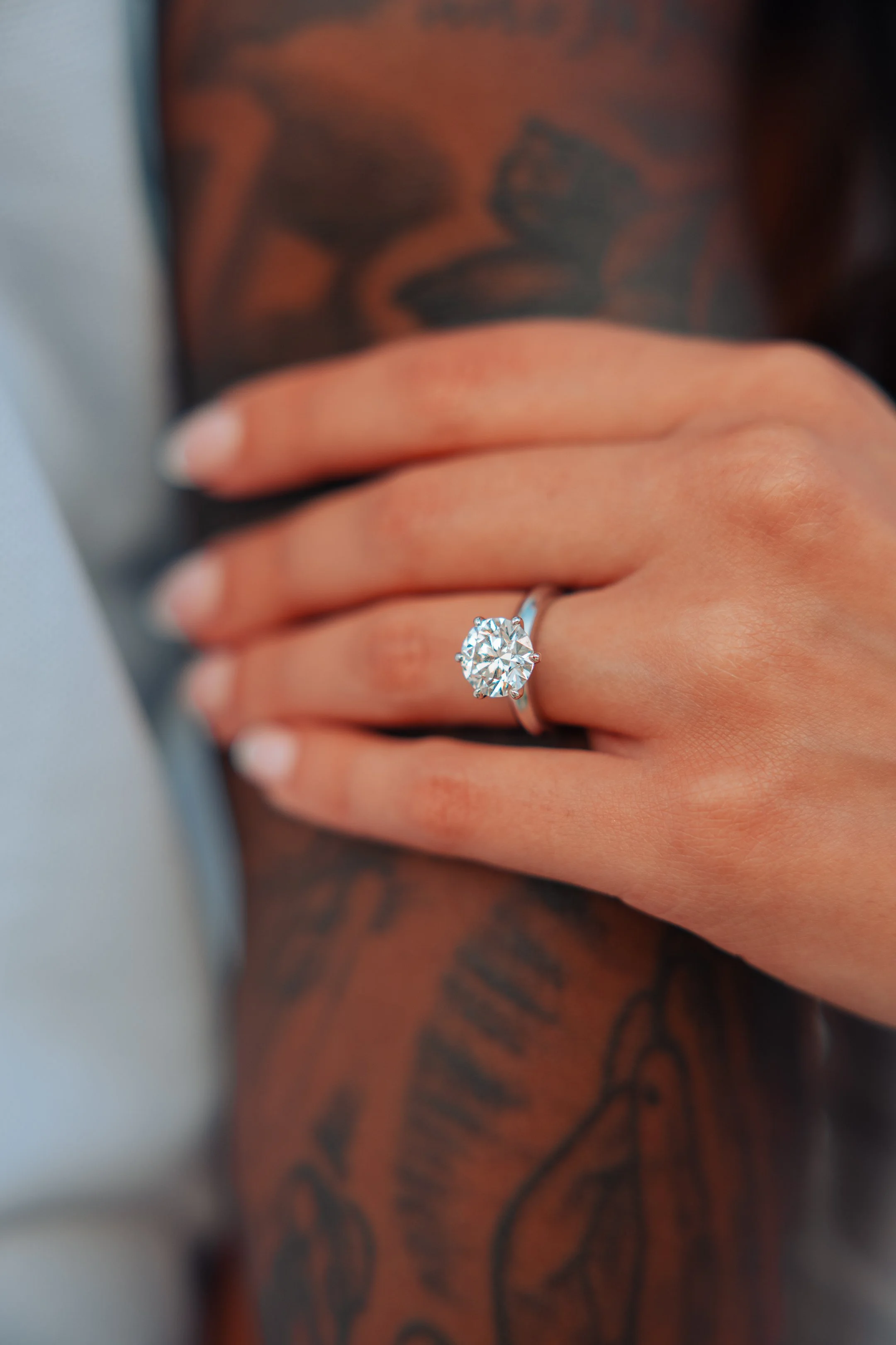 A close-up of a woman's hand with a diamond engagement ring on her ring finger, resting on a person's arm with tattoos.