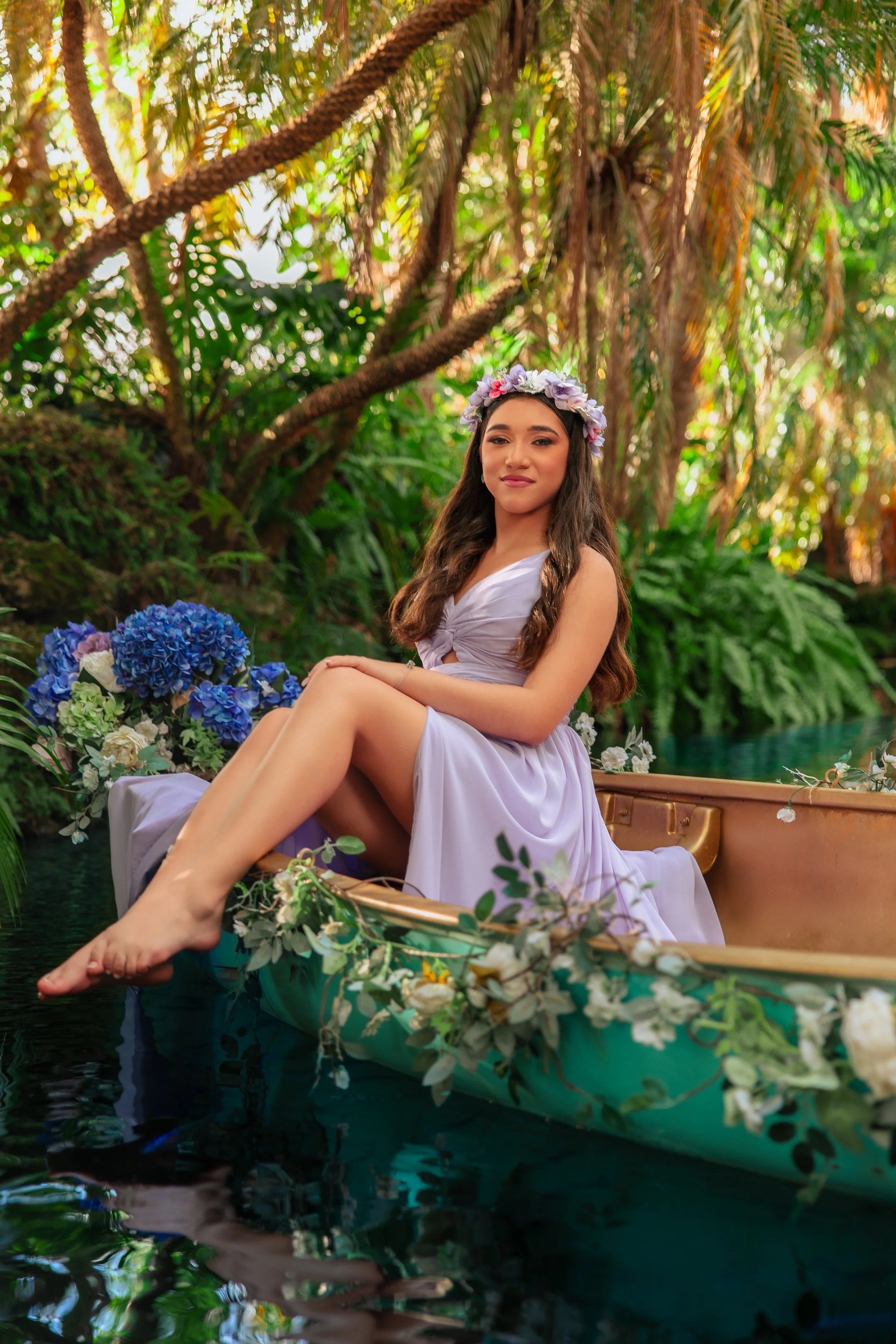 A young woman in a white dress with a flower crown, sitting on a decorated boat surrounded by lush greenery and flowers in a tropical setting.