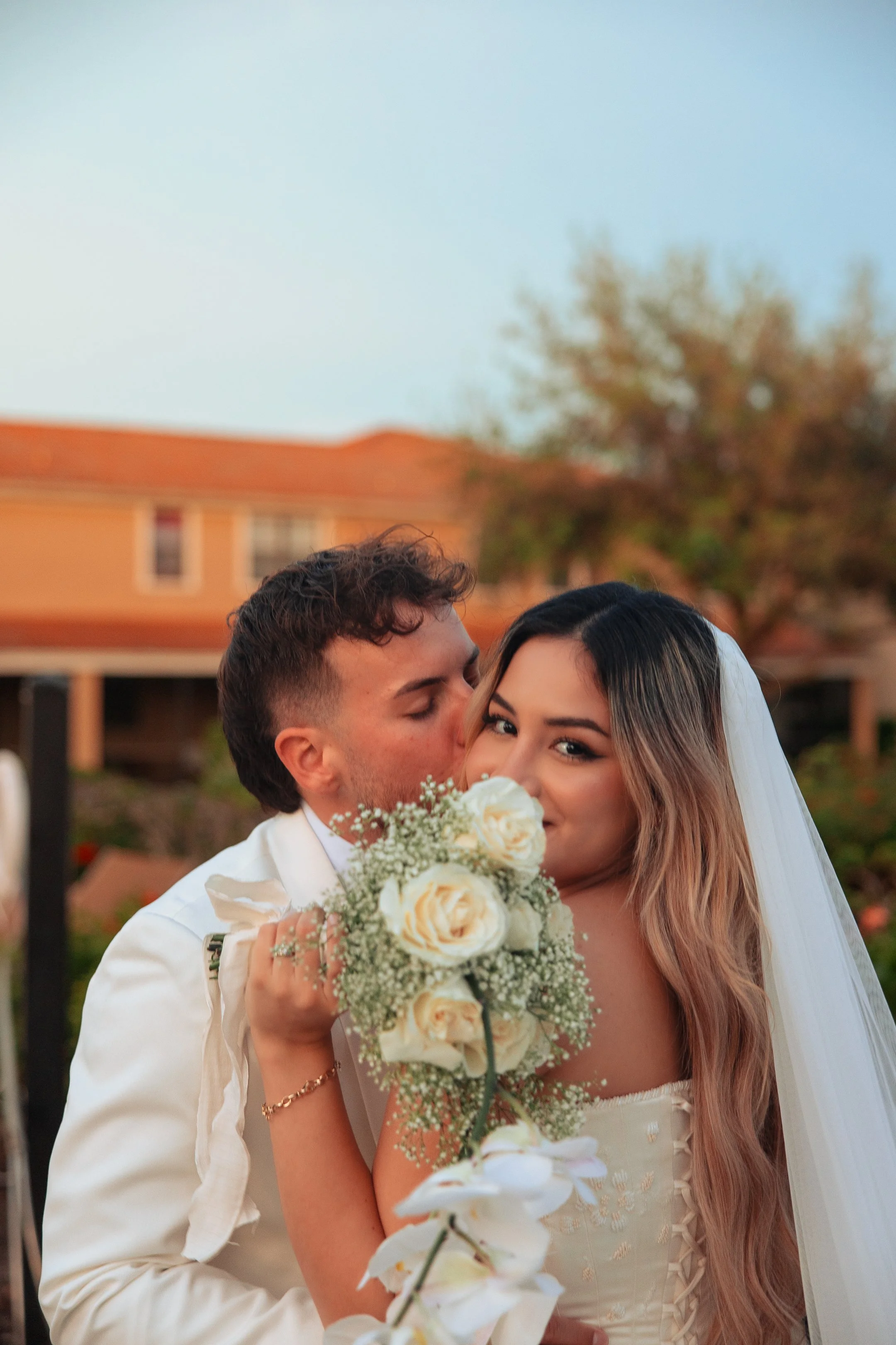 A bride and groom on their wedding day, with the groom kissing the bride's cheek as she holds a bouquet of white roses and baby's breath, outdoors during sunset.