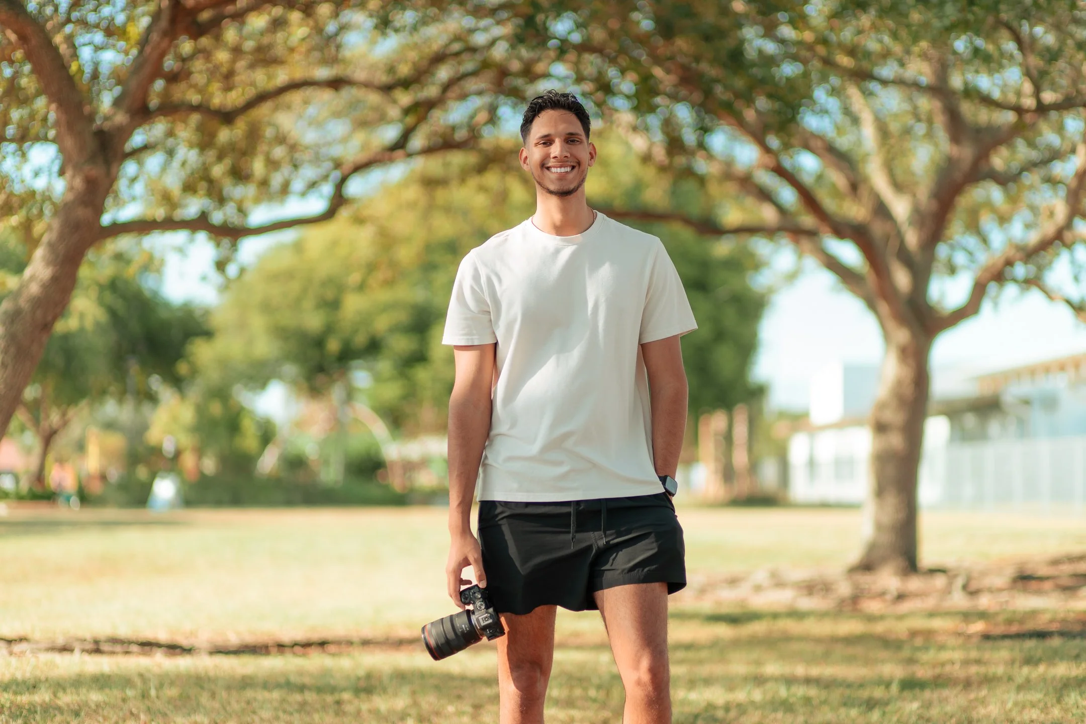 A young man standing outdoors in a park, holding a camera in his right hand and smiling at the camera. He is wearing a white t-shirt and black shorts, with trees and a bright sky in the background.