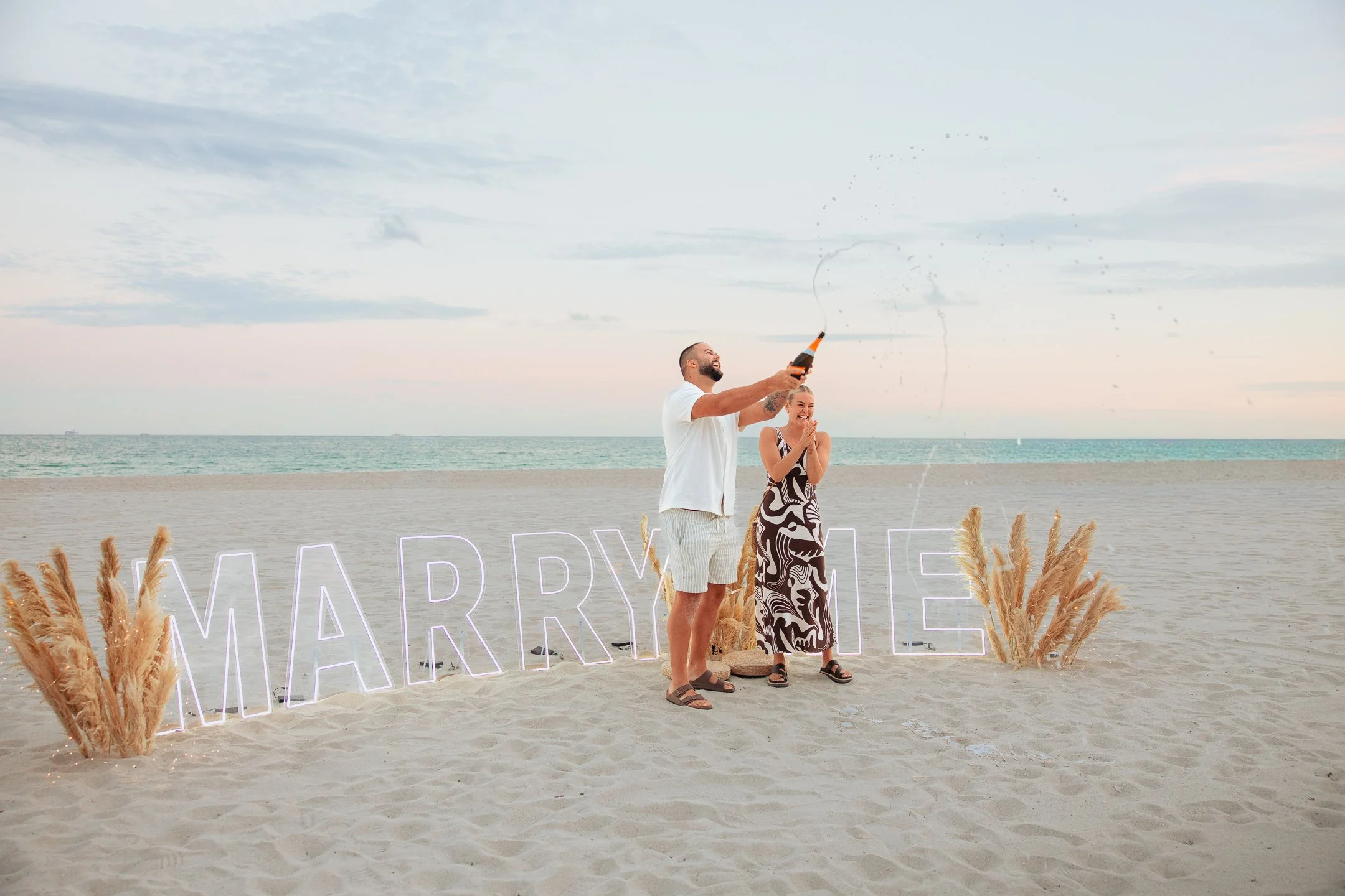 A couple celebrating marriage proposal on the beach with a large neon sign spelling 'MARRY ME' and pampas grass decorations.