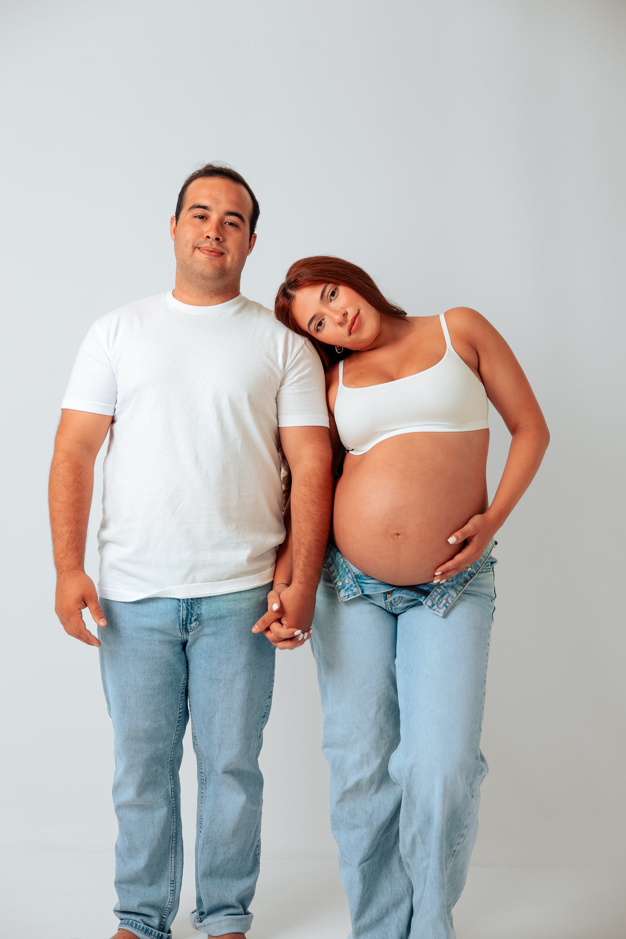 A pregnant woman with red hair leaning her head on a man’s shoulder, holding her belly, with a man standing beside her, all holding hands and looking at the camera against a plain light background.