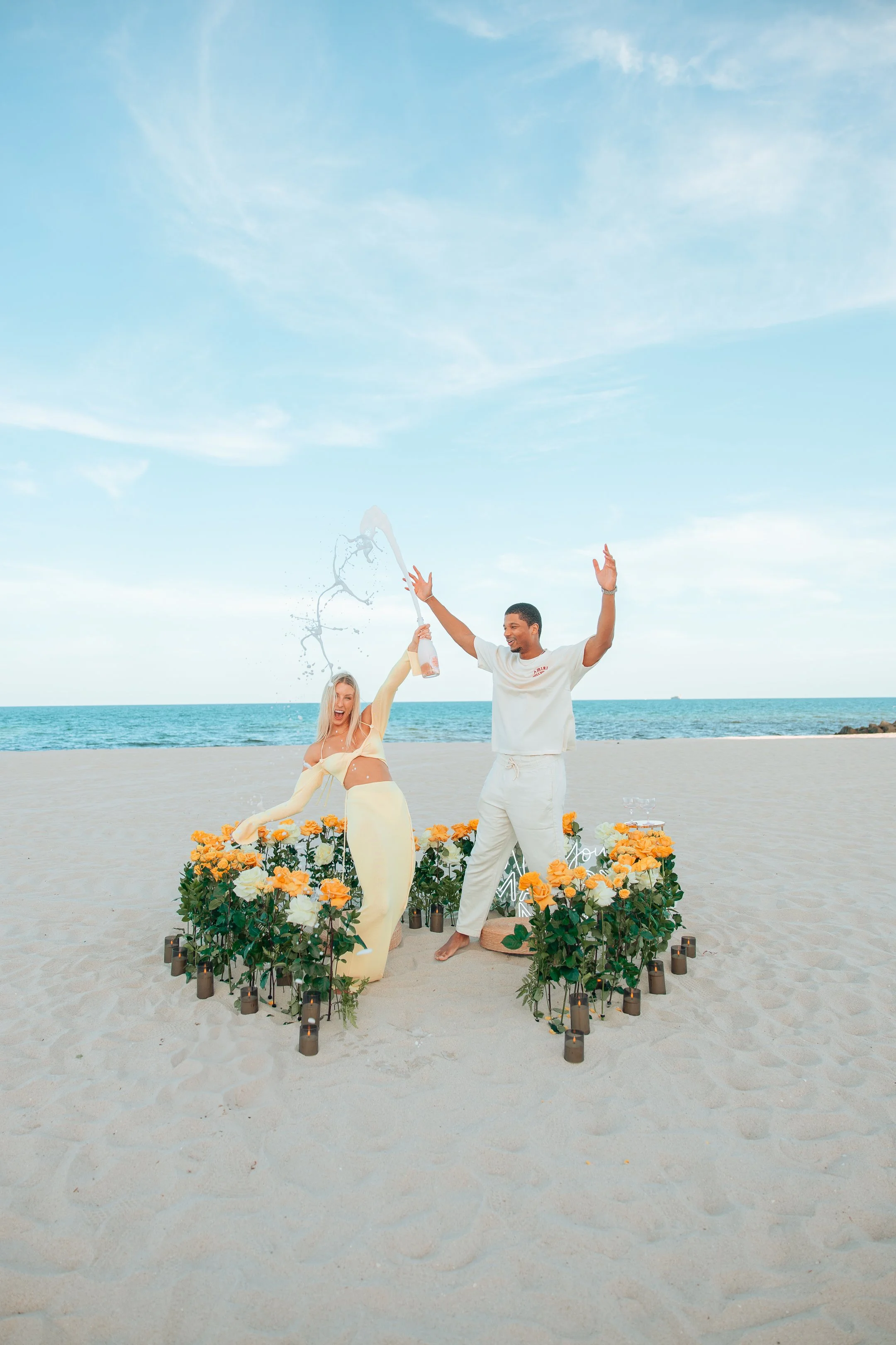 Couple celebrating on the beach with flowers and candles, one holding a champagne bottle spraying it, with a blue sky and ocean in the background.