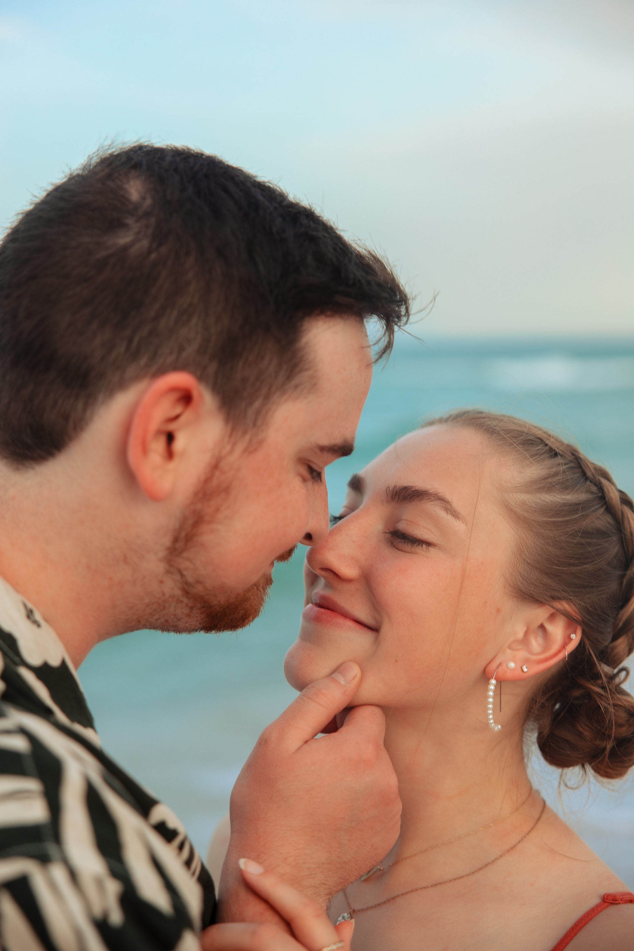 A couple touching foreheads and smiling with eyes closed on the beach during daytime, ocean in the background.