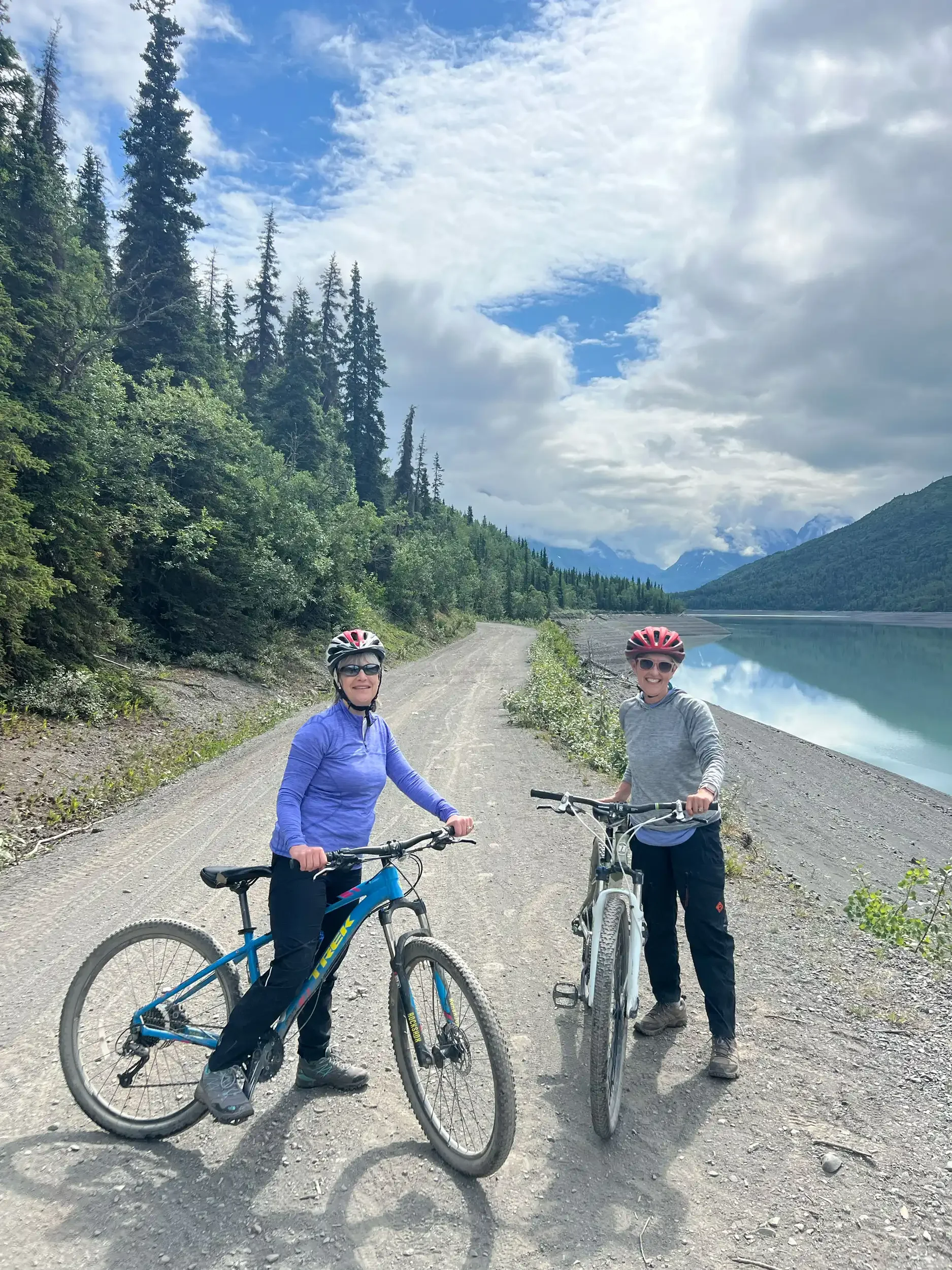 Two women with helmets and sunglasses standing with mountain bikes on a dirt trail beside a lake