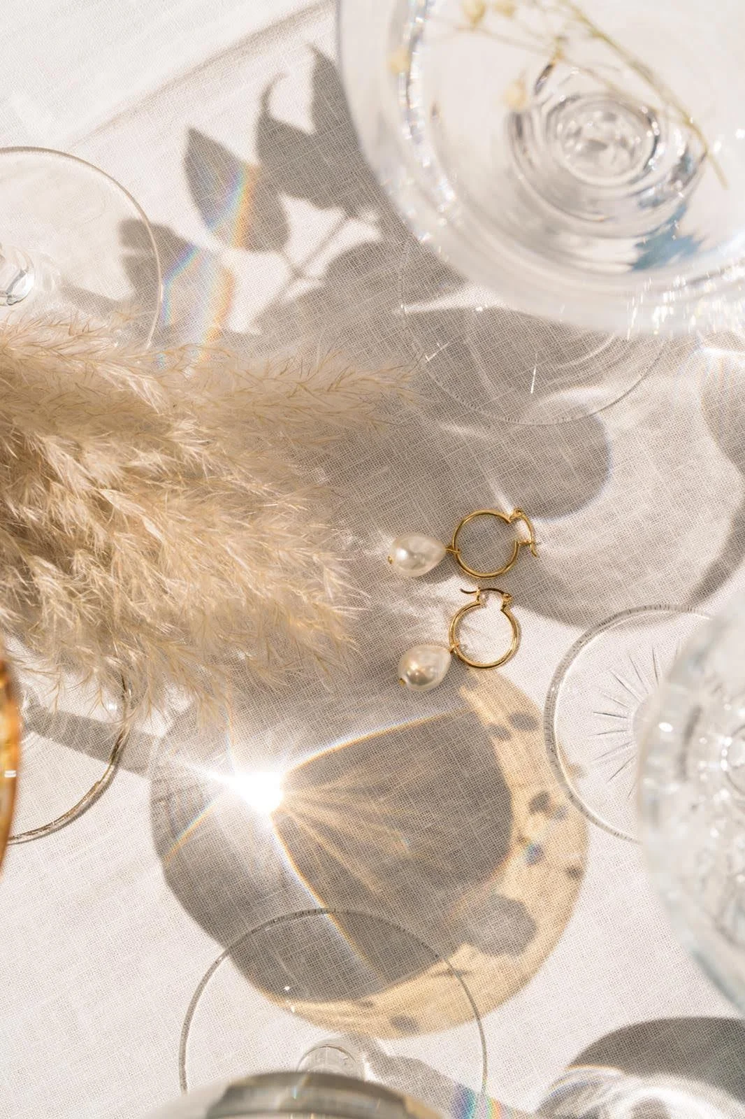 Overhead view of pampas grass, gold hoop earrings with pearls, and shadows of glassware on a light tablecloth.