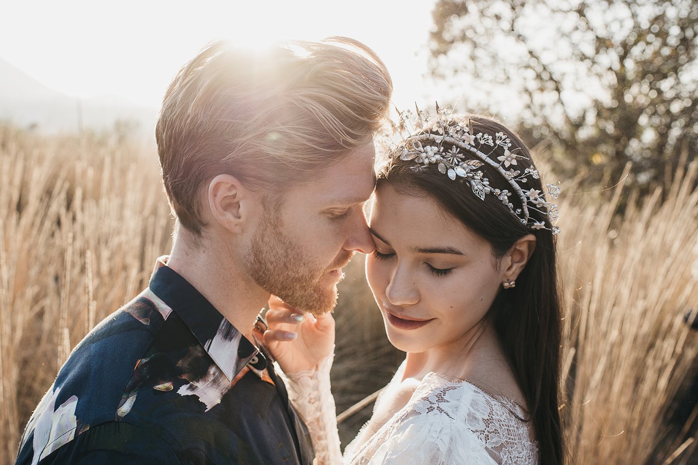 Close-up of a couple embracing outdoors, with a woman wearing a floral headpiece and a man in a patterned shirt, surrounded by tall grass with sunlight in the background.