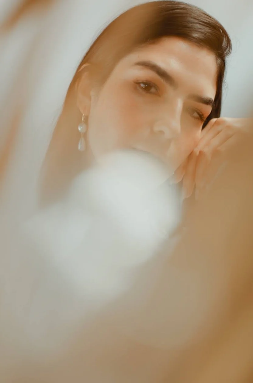 Soft-focused portrait of a woman with long dark hair, resting her chin on her hand, wearing pearl earrings.