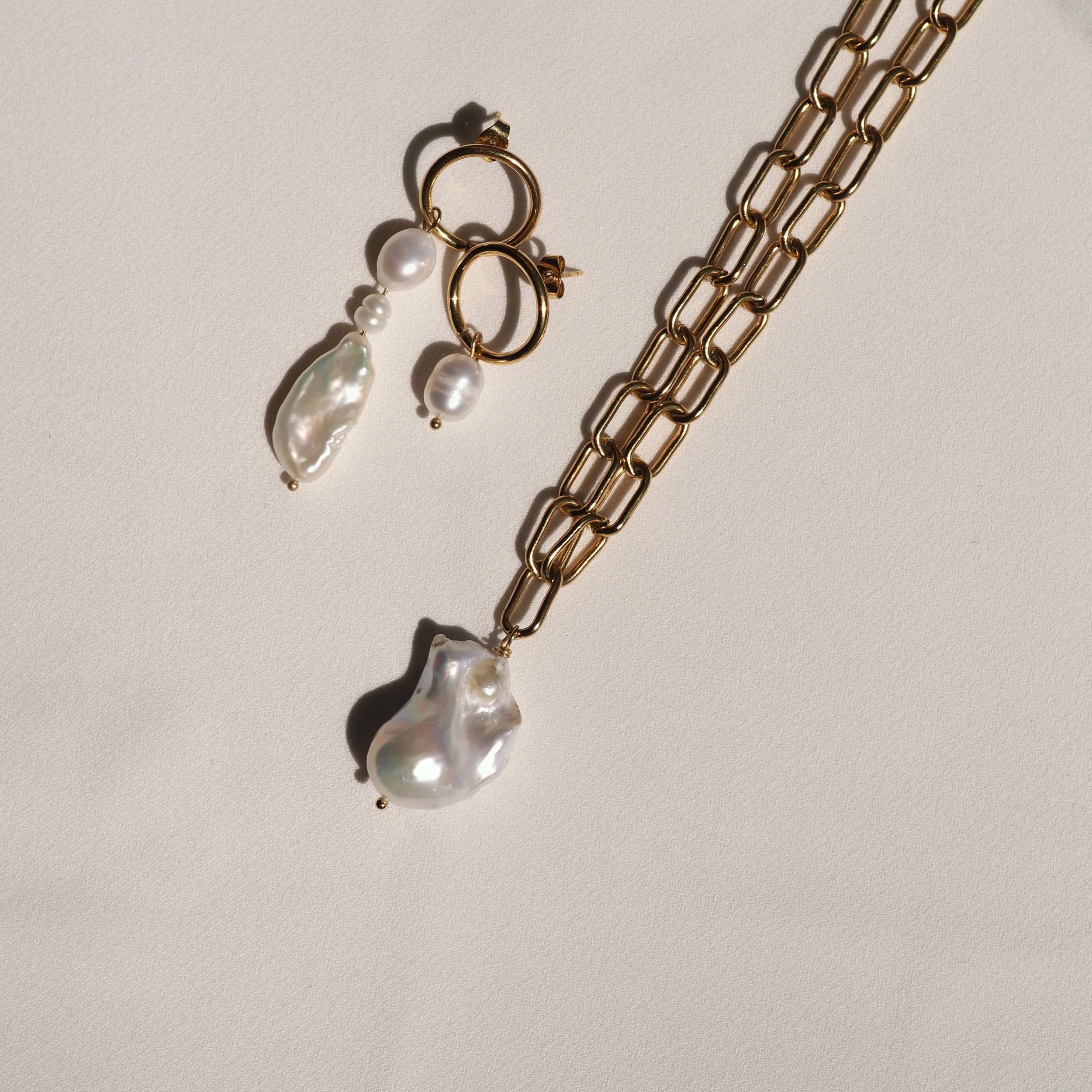 Gold chain necklace with large baroque pearl pendant and matching gold hoop earrings with pearls against a light background.