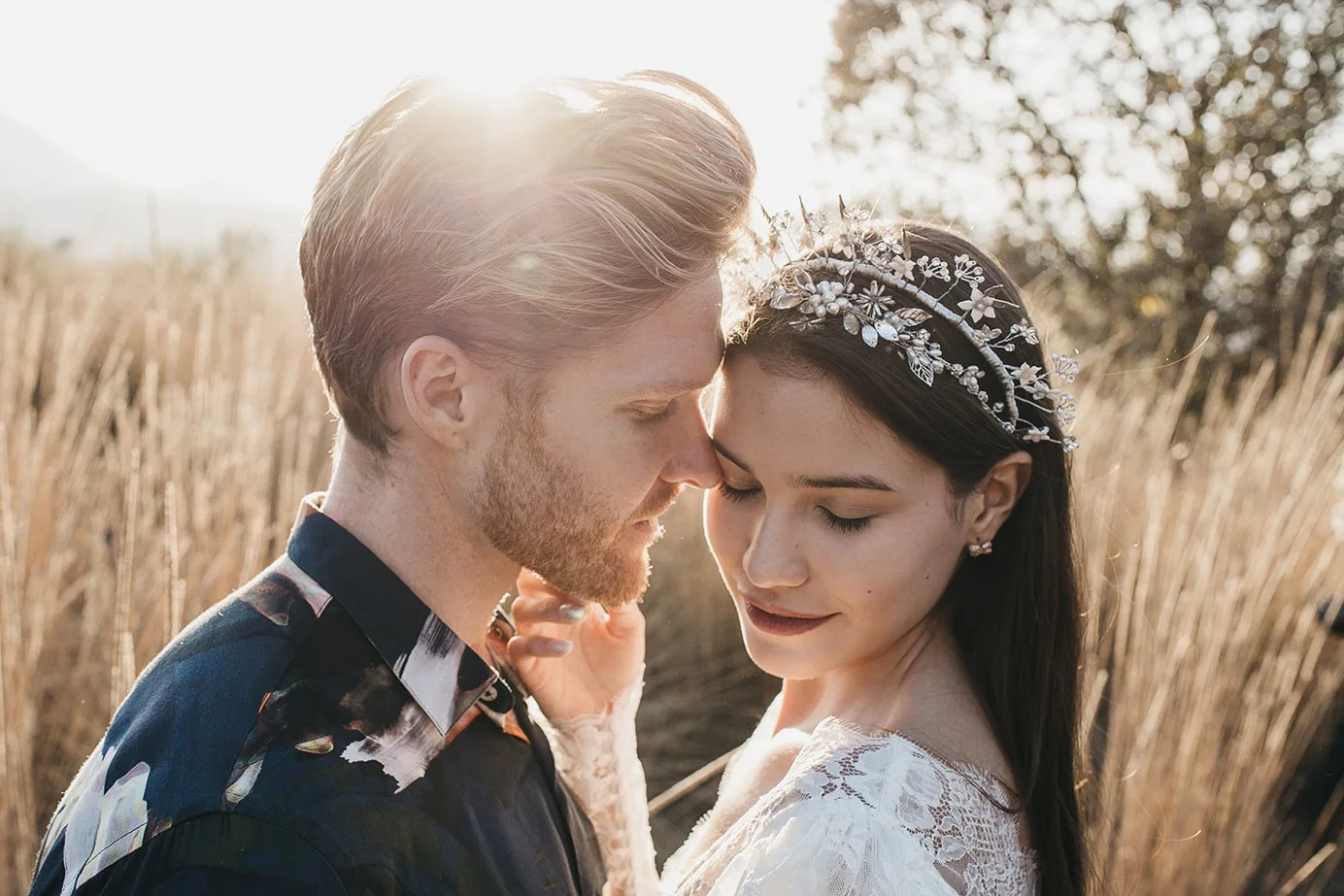 A couple embraces outdoors with a sunlit backdrop, the woman wearing a floral headpiece and lace attire.
