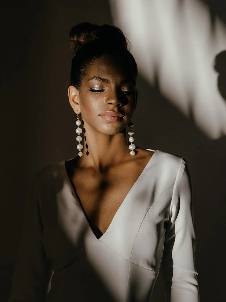 A woman in a white dress with long pearl earrings, eyes closed, standing in dramatic lighting.