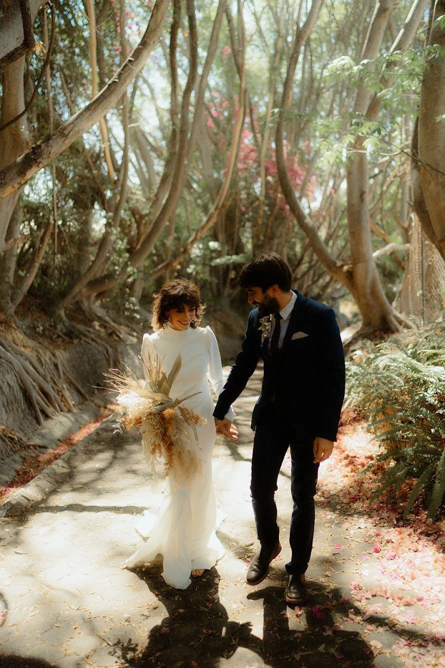 Bride and groom walking hand in hand through a sunlit forest path, with the bride holding a bouquet of dried flowers.