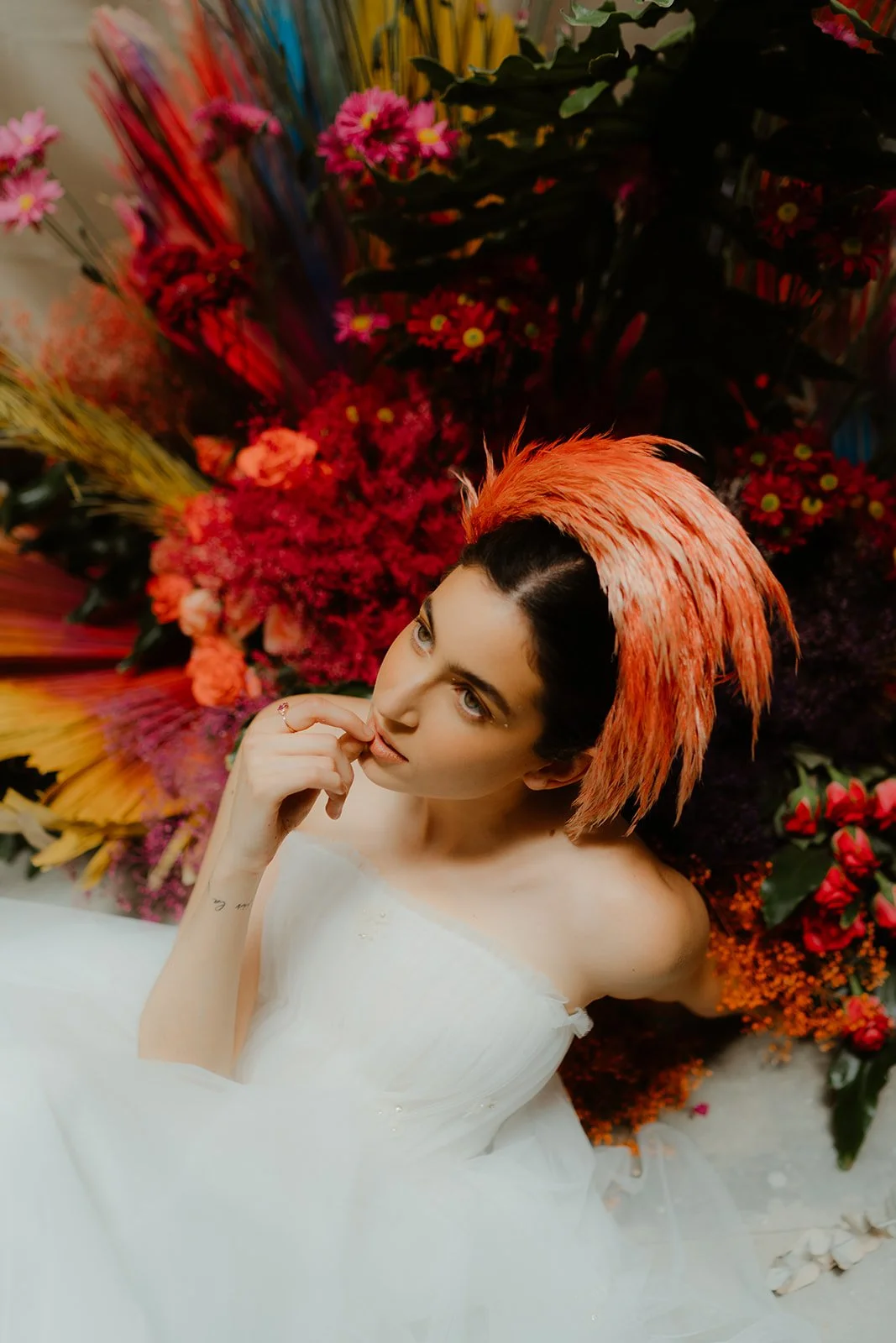 Woman in white dress with orange feathered headpiece, surrounded by vibrant, colorful flowers.