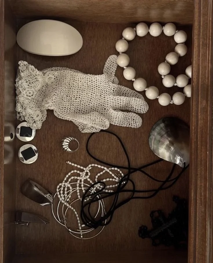A drawer containing a lace glove, white beaded necklace, metal rings, dome-shaped object, seashell, pearl necklace, black cord, and various small accessories.