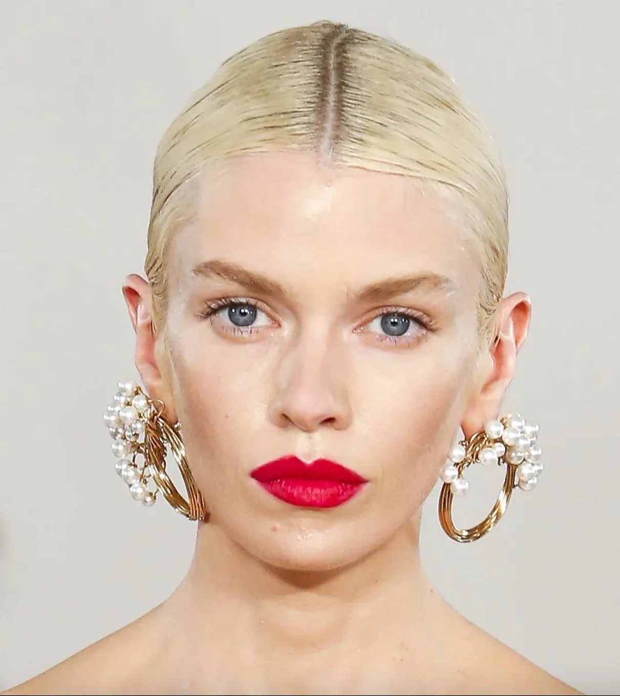 Person with slicked-back blonde hair, wearing large gold hoop earrings adorned with pearls, and bright red lipstick.