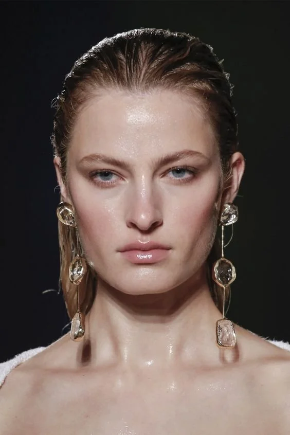 Model wearing unique long earrings and dewy makeup on a runway.