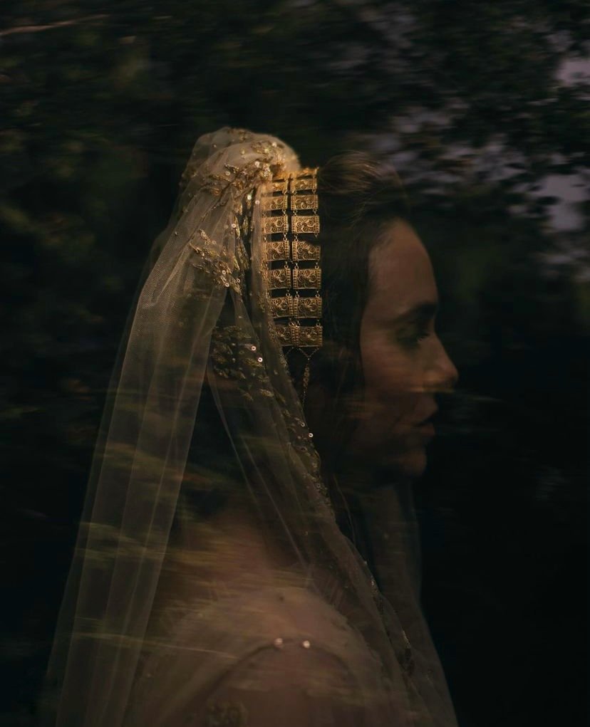 Profile of a woman wearing a dark, intricately detailed veil and headpiece against a blurred natural background.