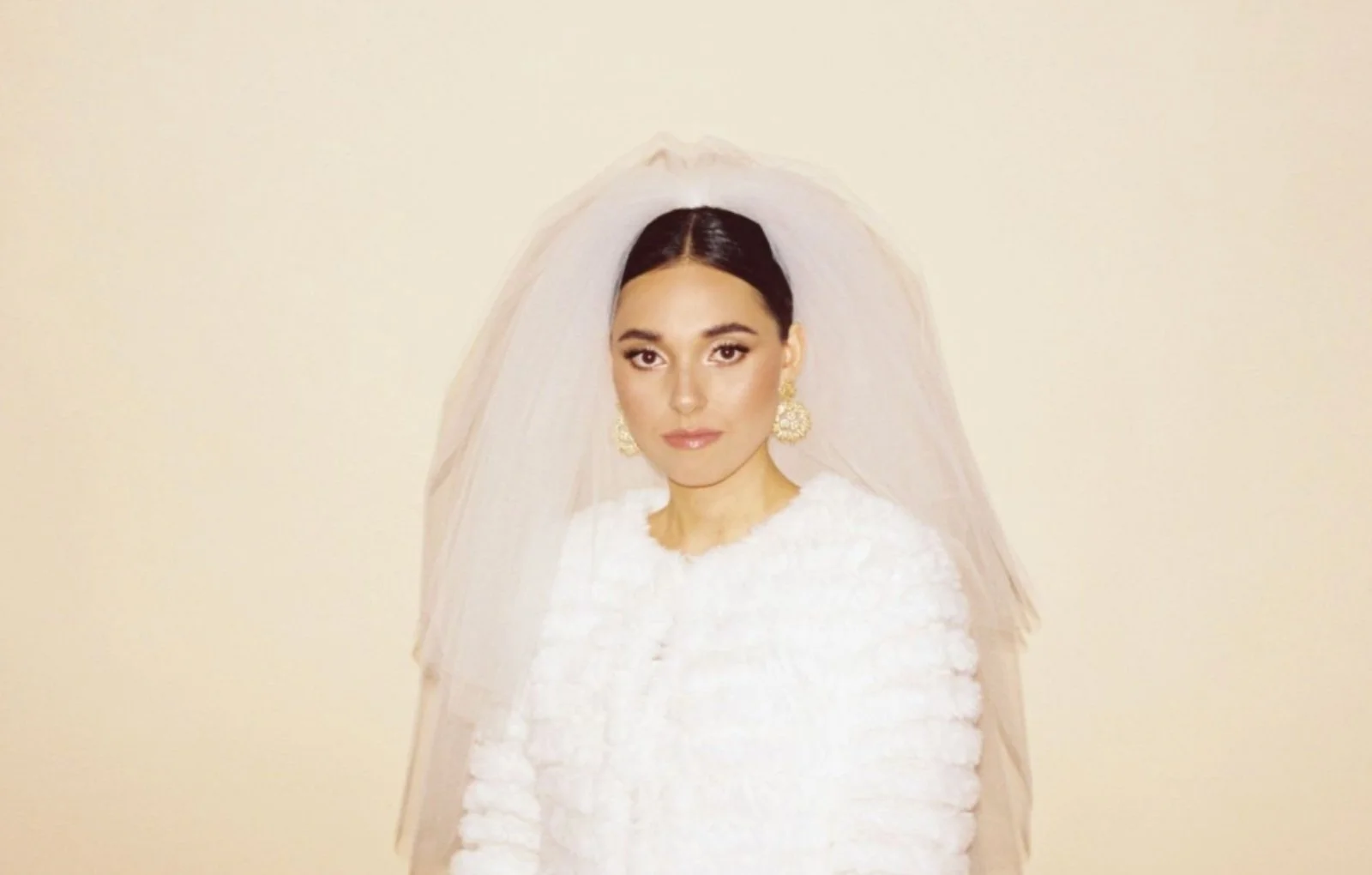 Woman in bridal outfit with veil and ornate earrings against a light background.